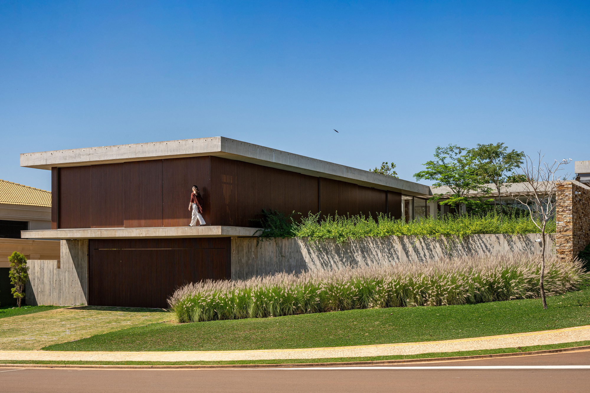 Street view of the dark timber-clad upper volume with concrete soffit above planted grasses and lawn