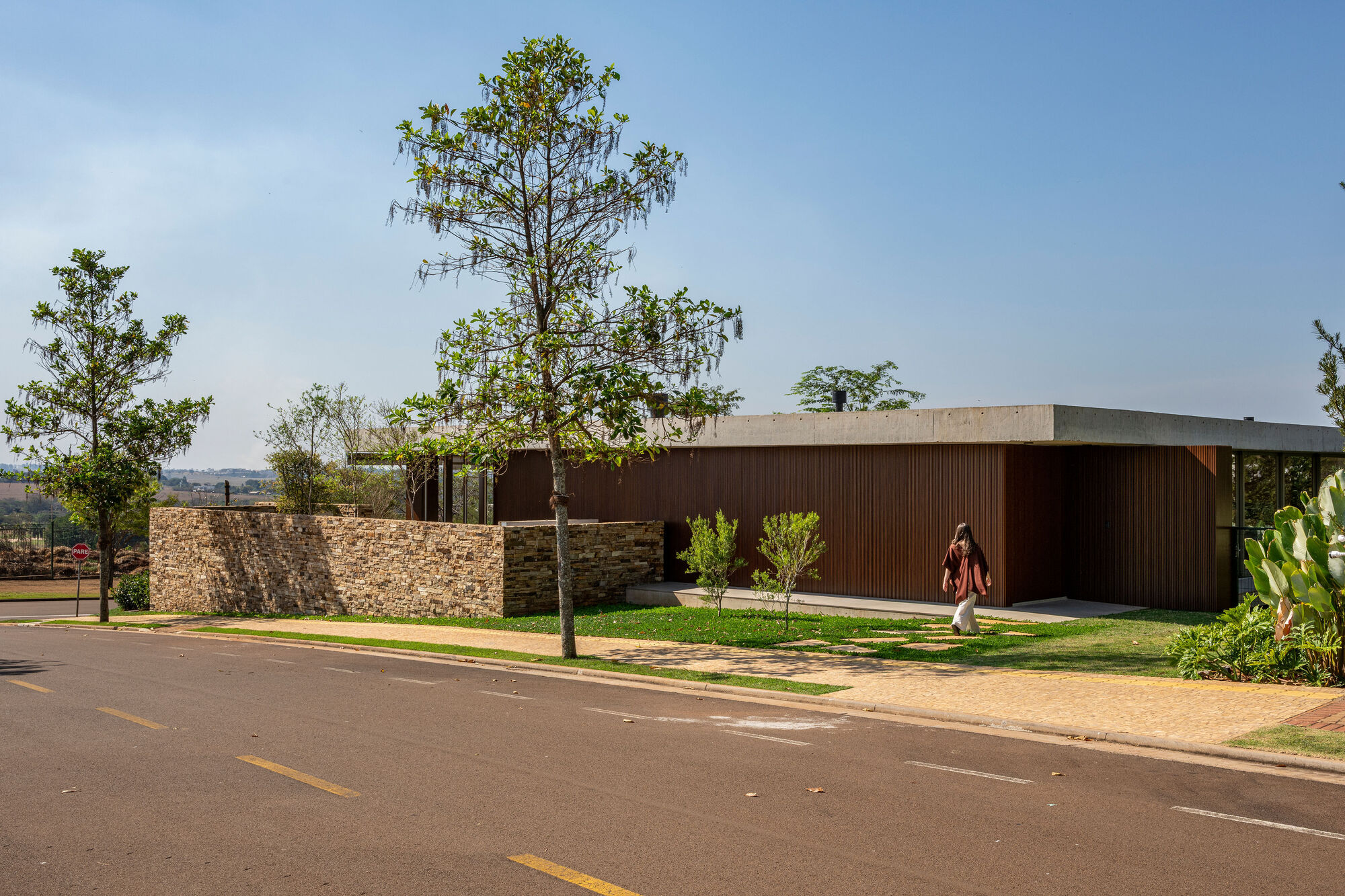 Stone perimeter wall and dark timber facade from the street with scattered trees under clear sky