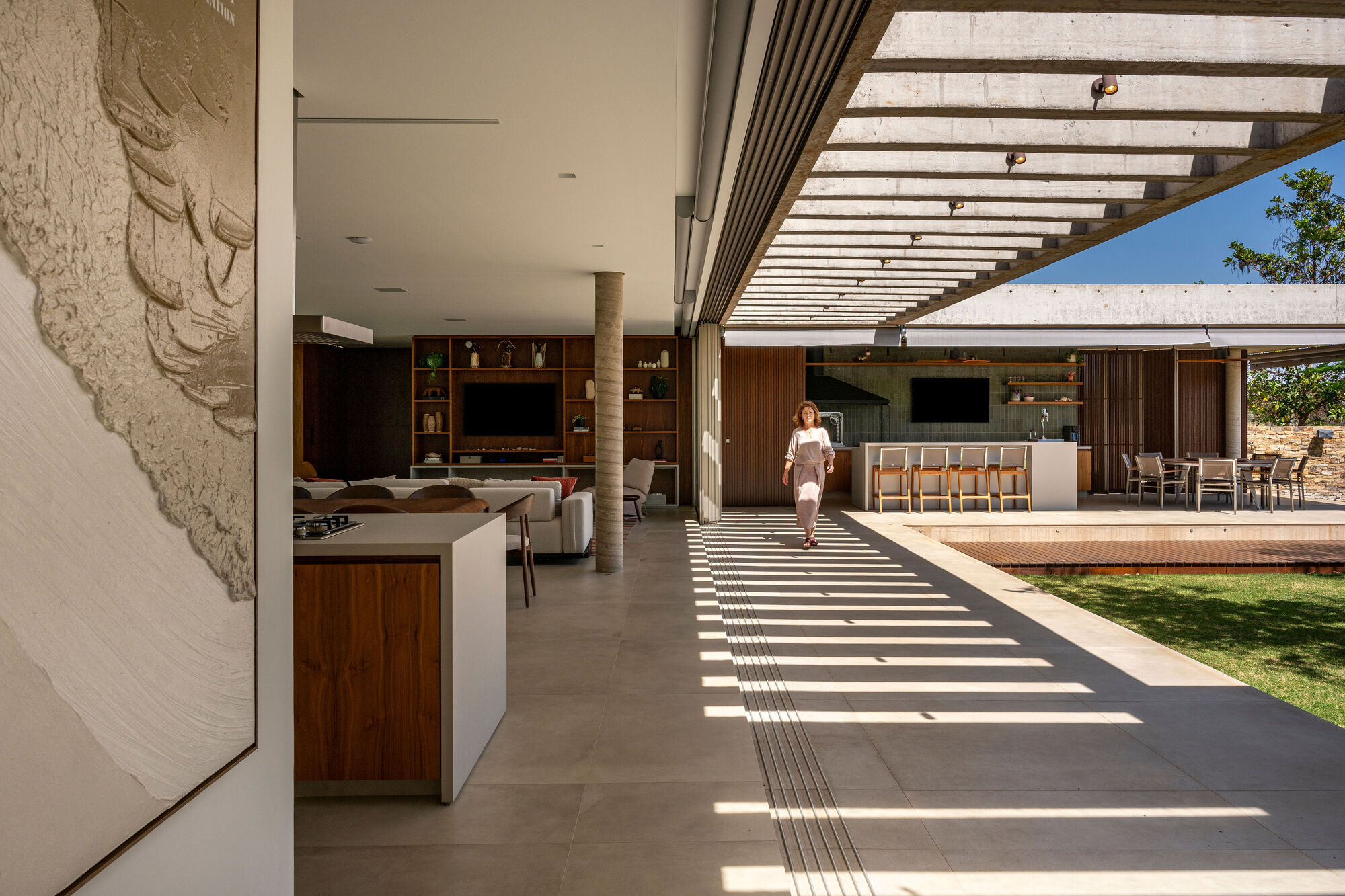 Open living pavilion with slatted pergola casting shadows across the floor as a figure walks through