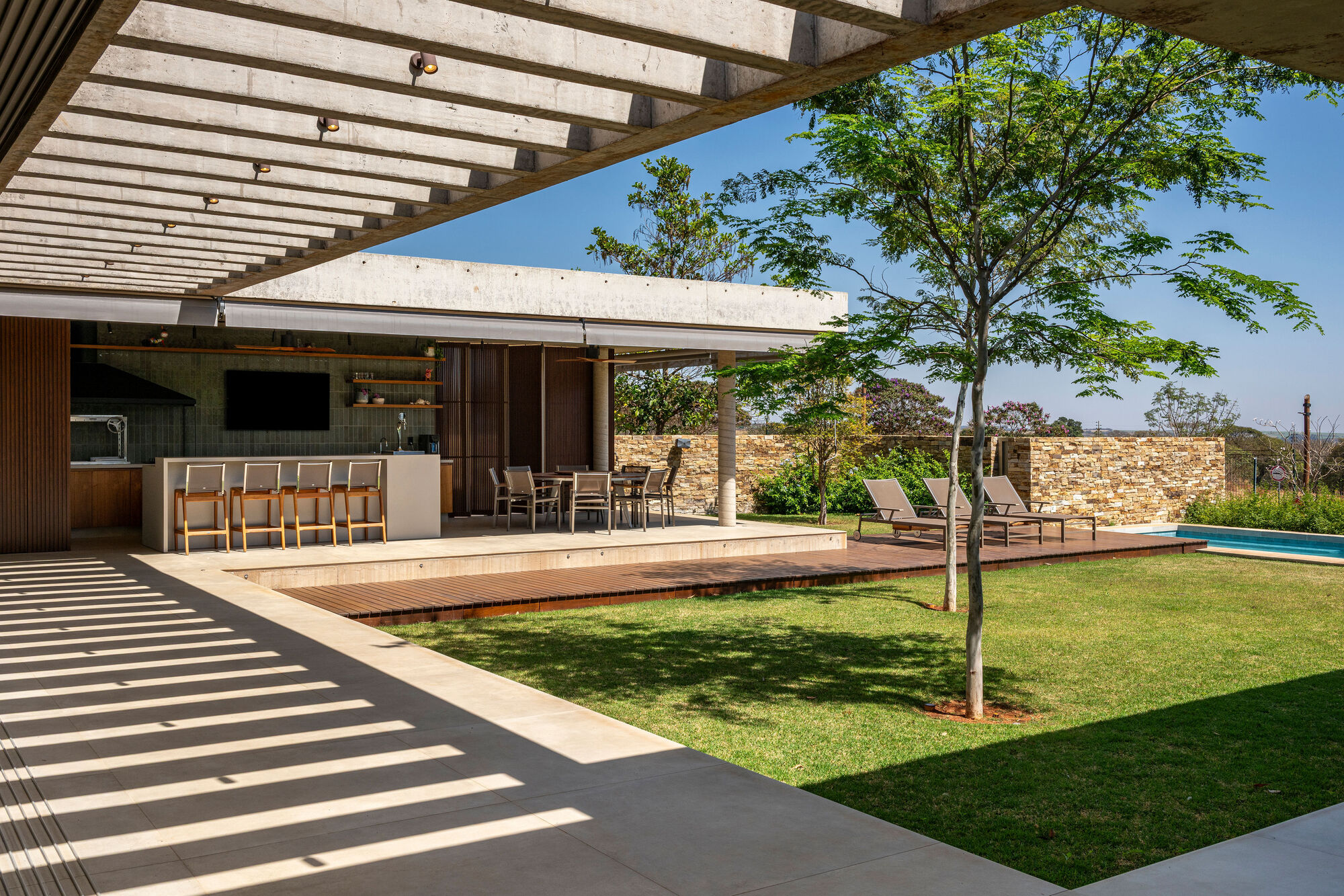 Covered terrace with concrete pergola beams casting shadows onto the lawn and outdoor kitchen