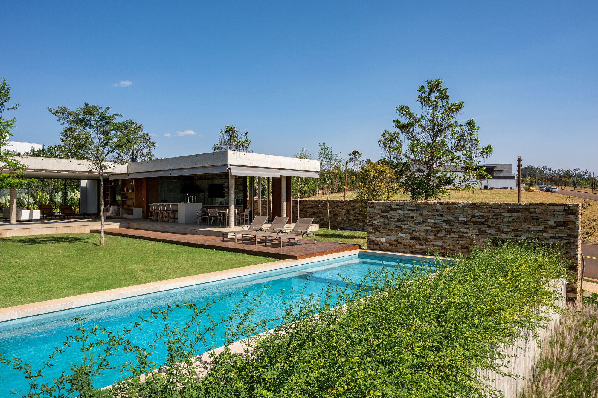 Lap pool edged with planted beds and a stone wall under a clear afternoon sky