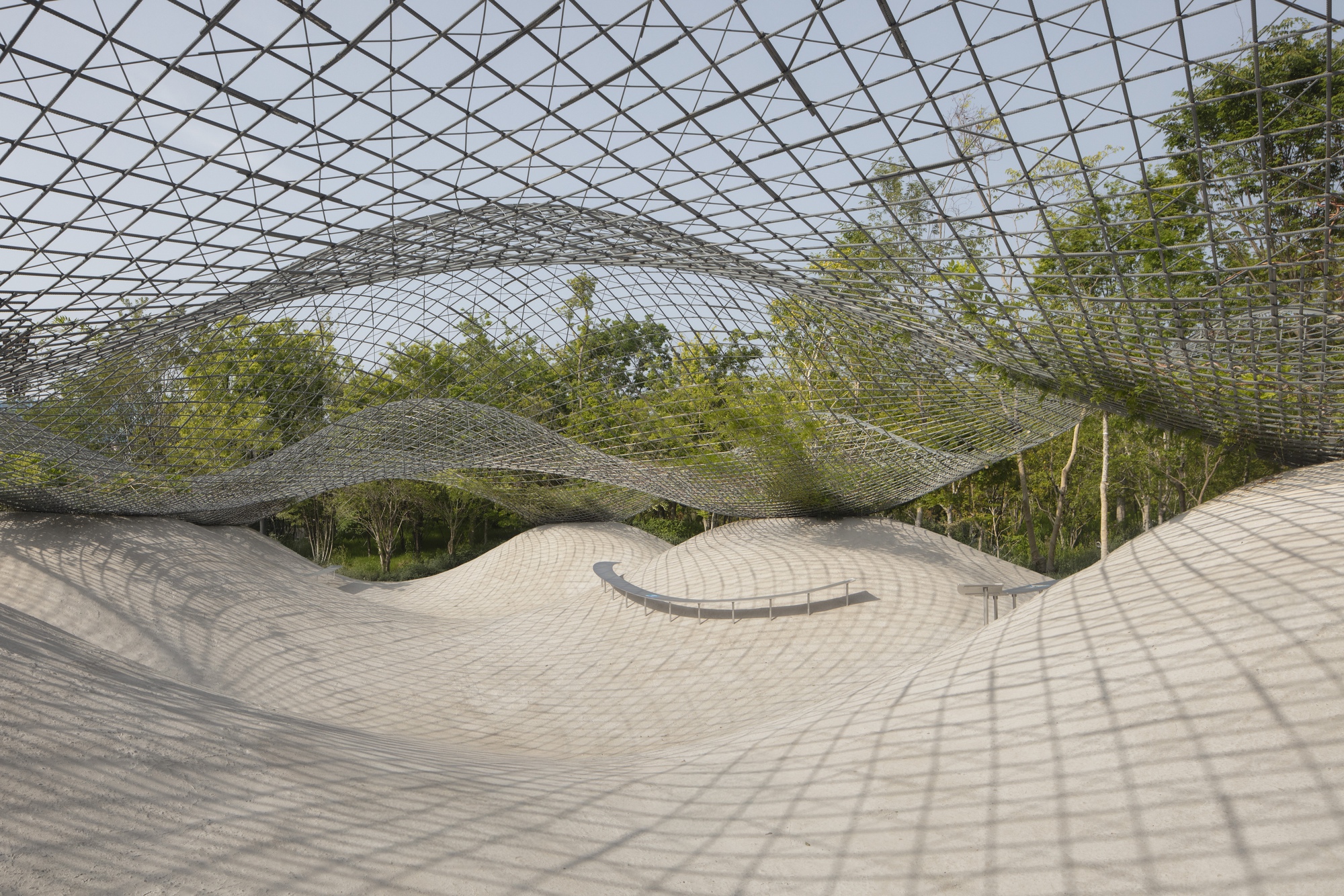 Undulating paved courtyard beneath a diagrid metal canopy with dappled shadows from overhead trees