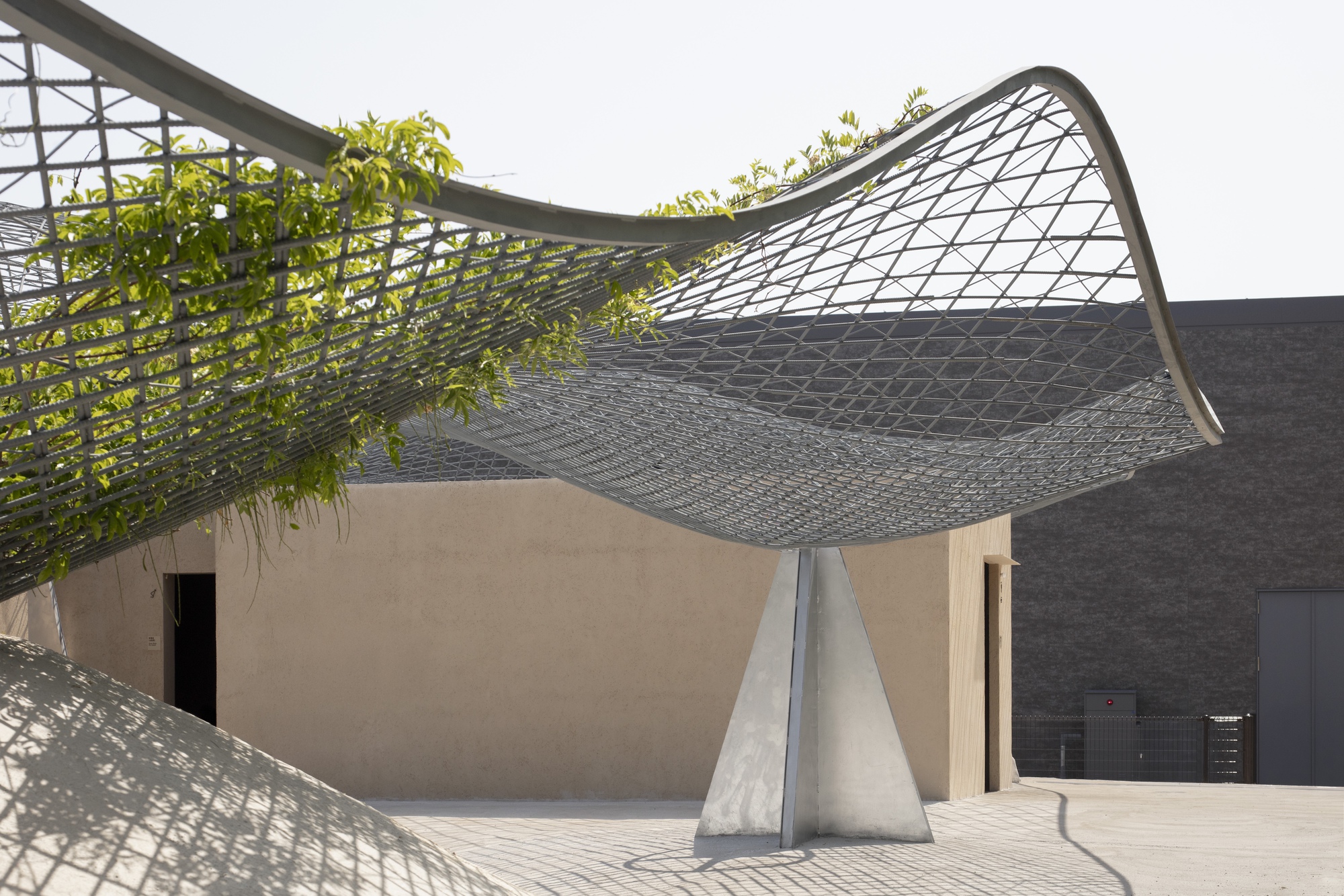 Curved metal mesh canopy rising from concrete supports over a courtyard with planted greenery