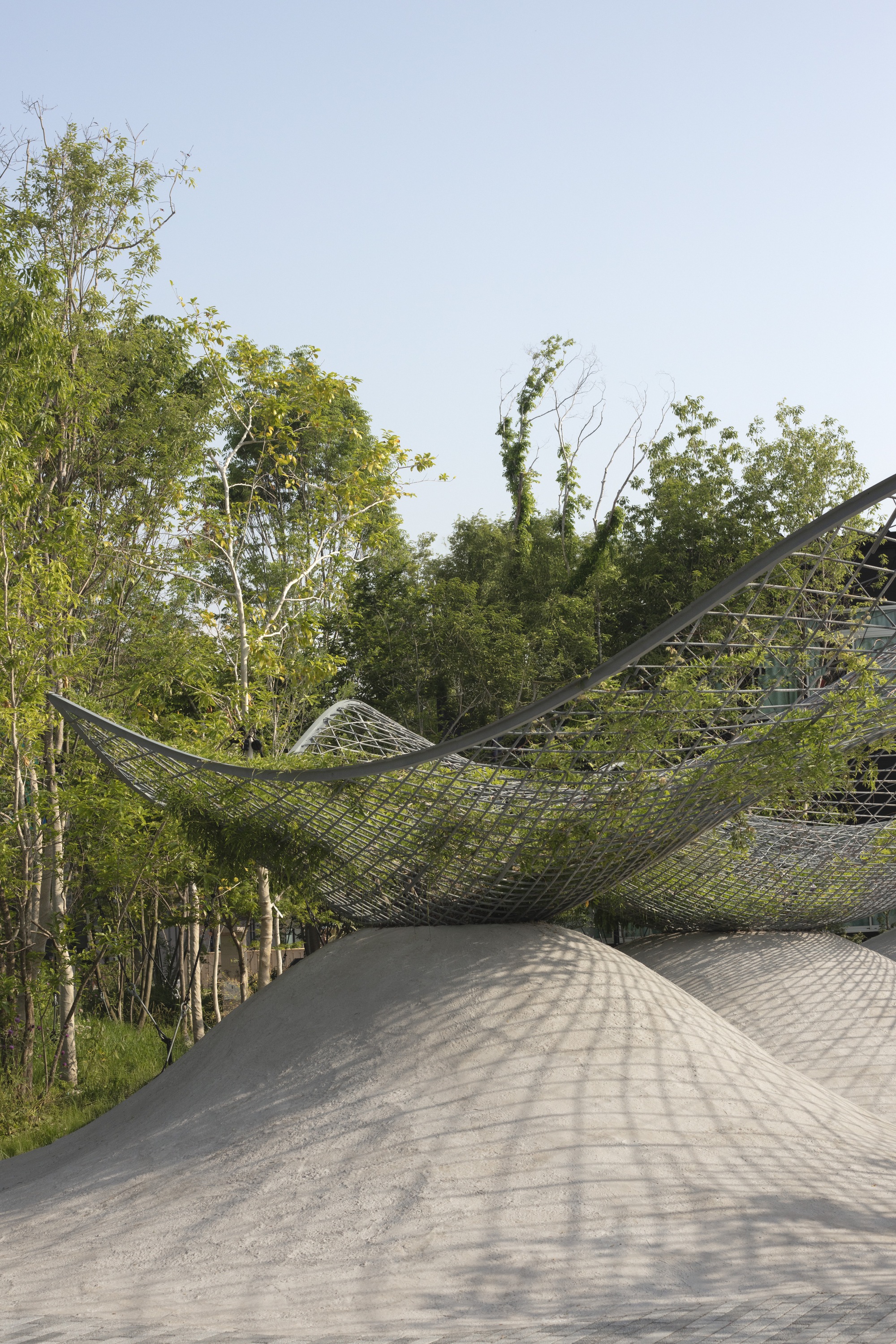 Curved concrete roof beneath a tensile steel mesh canopy suspended above leafy birch trees