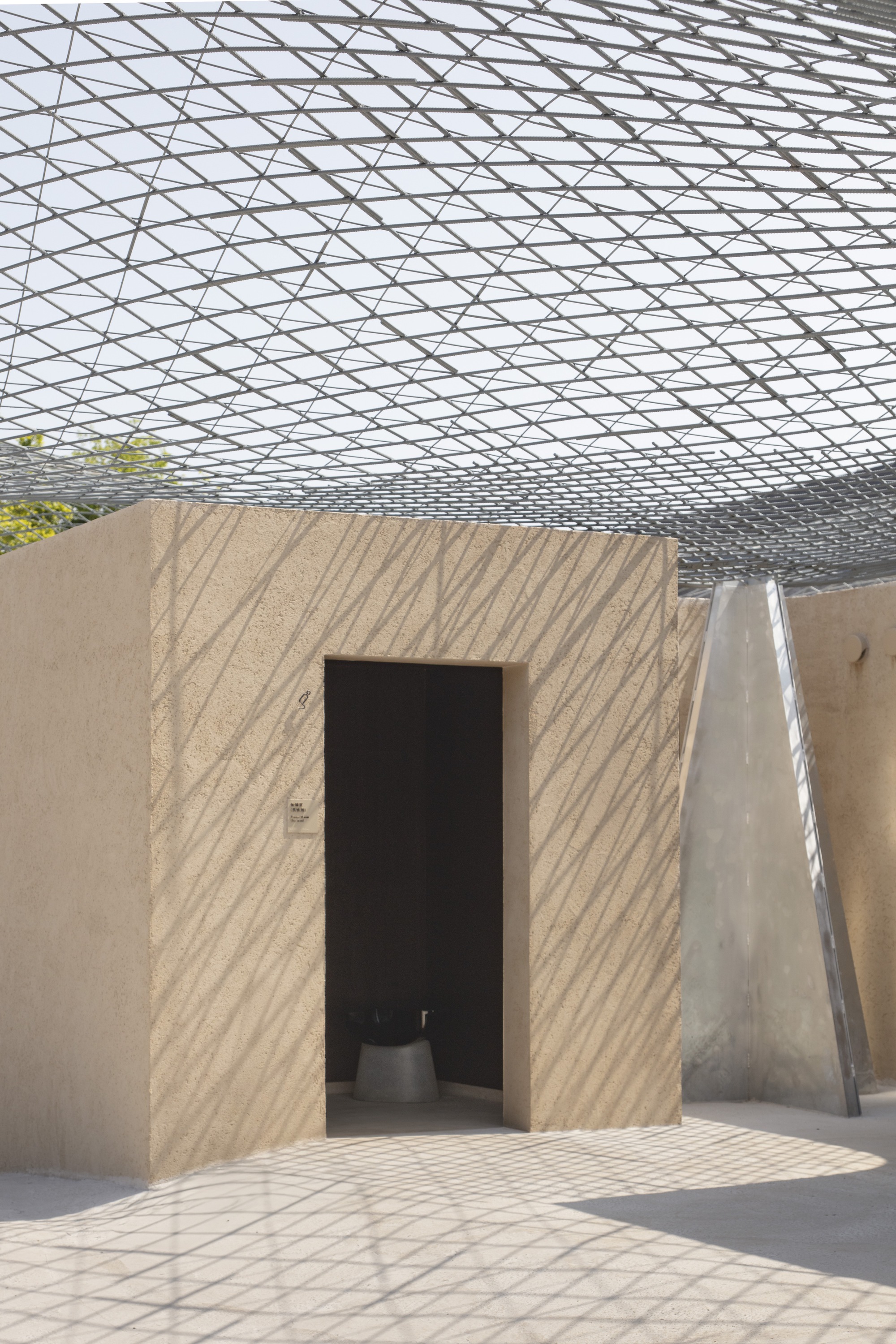 Textured rammed earth doorway beneath the geometric shadow pattern cast by the overhead mesh structure