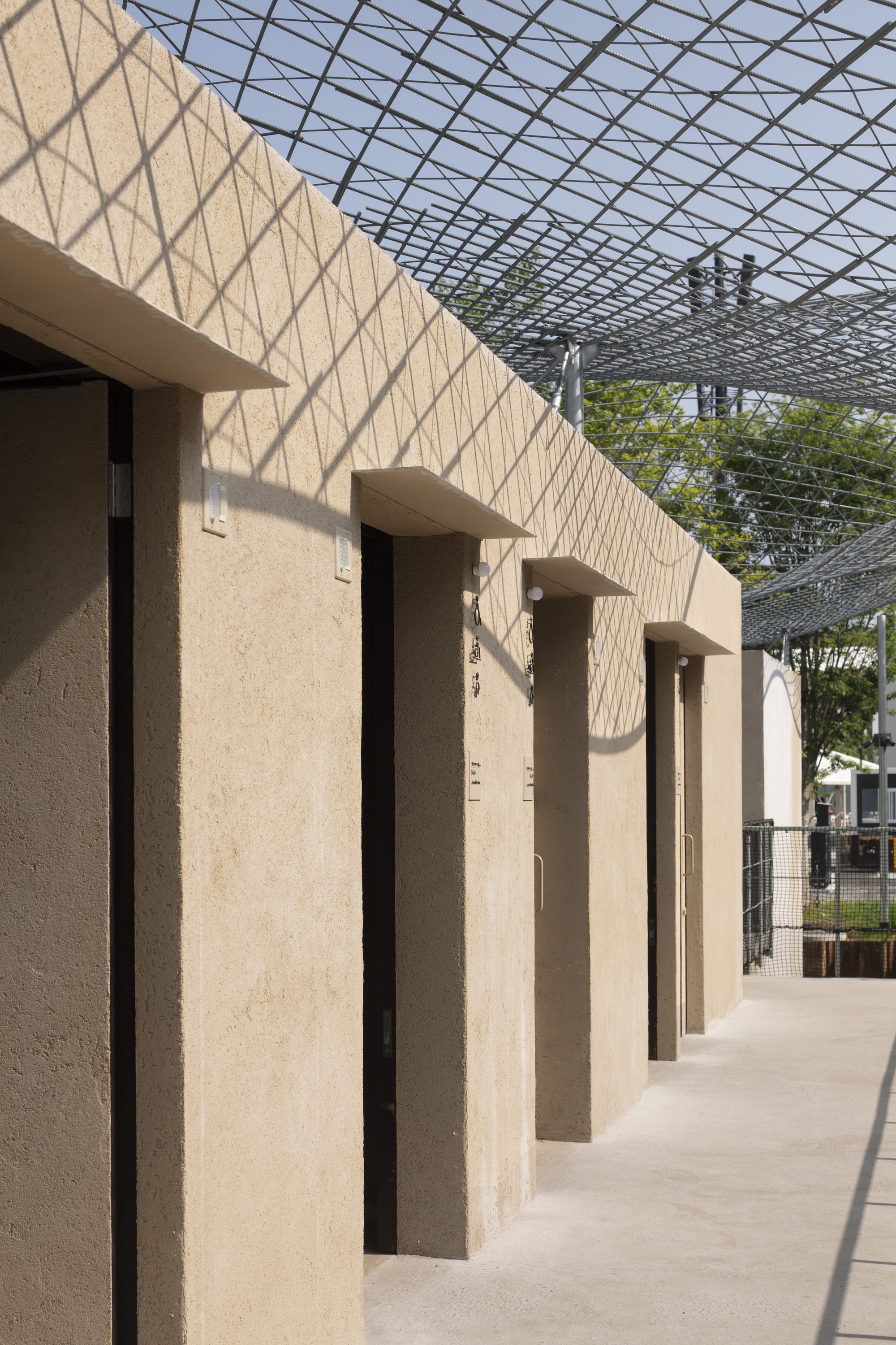 Row of vertical recesses in the rammed earth facade creating rhythmic shadows under the canopy