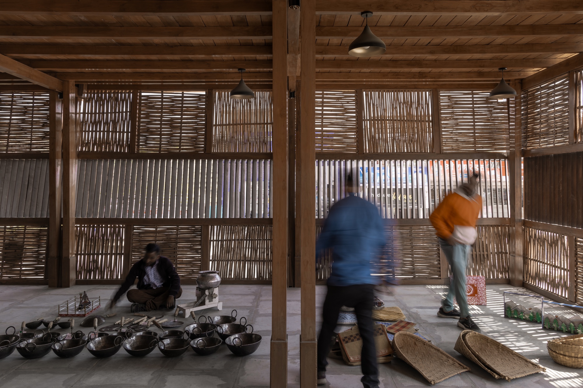 Covered workshop space with timber columns and woven screens where blurred figures arrange ceramic vessels on floor