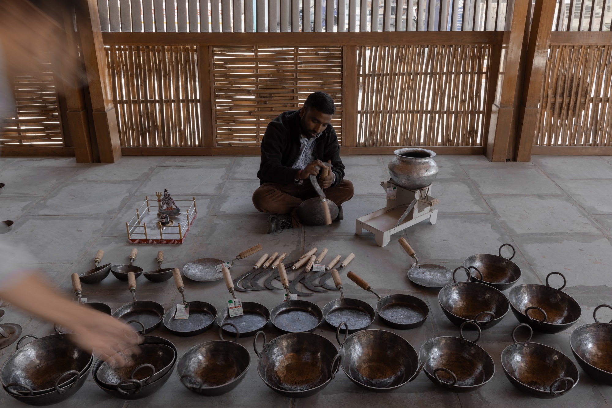 Seated artisan working among rows of metal cookware and handtools beneath slatted timber screen wall