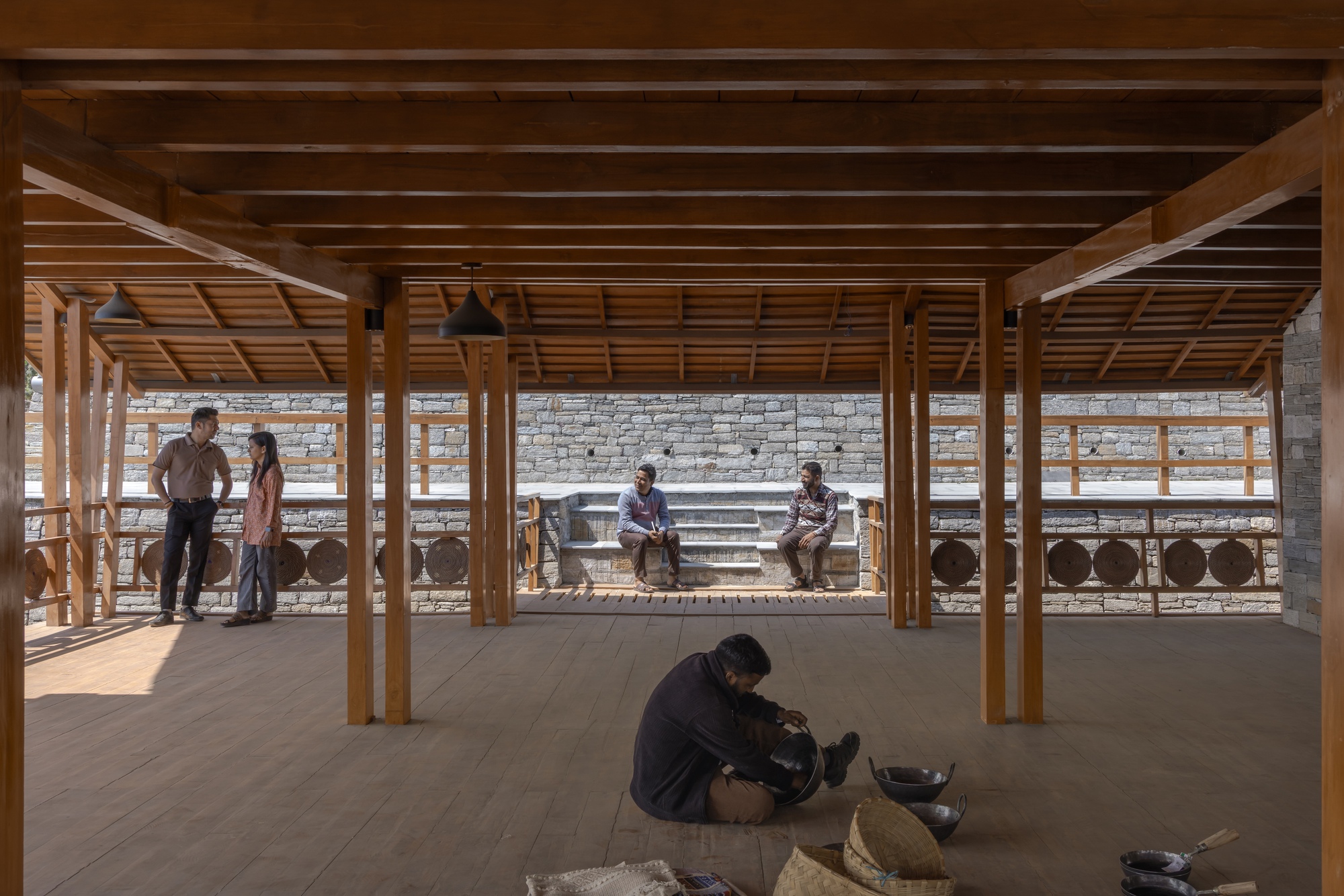 Covered timber pavilion with people gathered and seated along a stone wall with circular openings