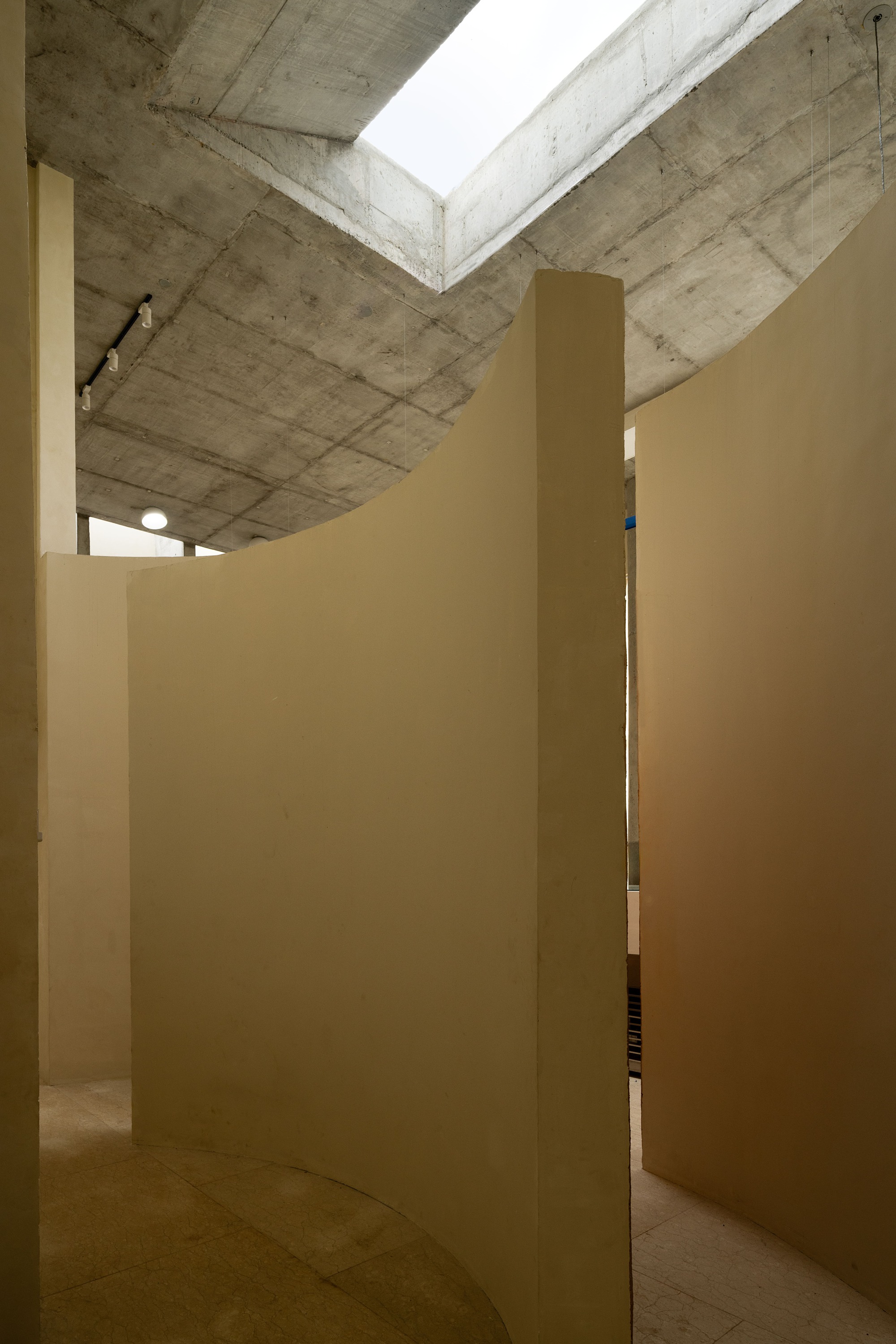 Narrow passage between cream-colored walls leading to glimpse of cork counter under concrete beam with skylight