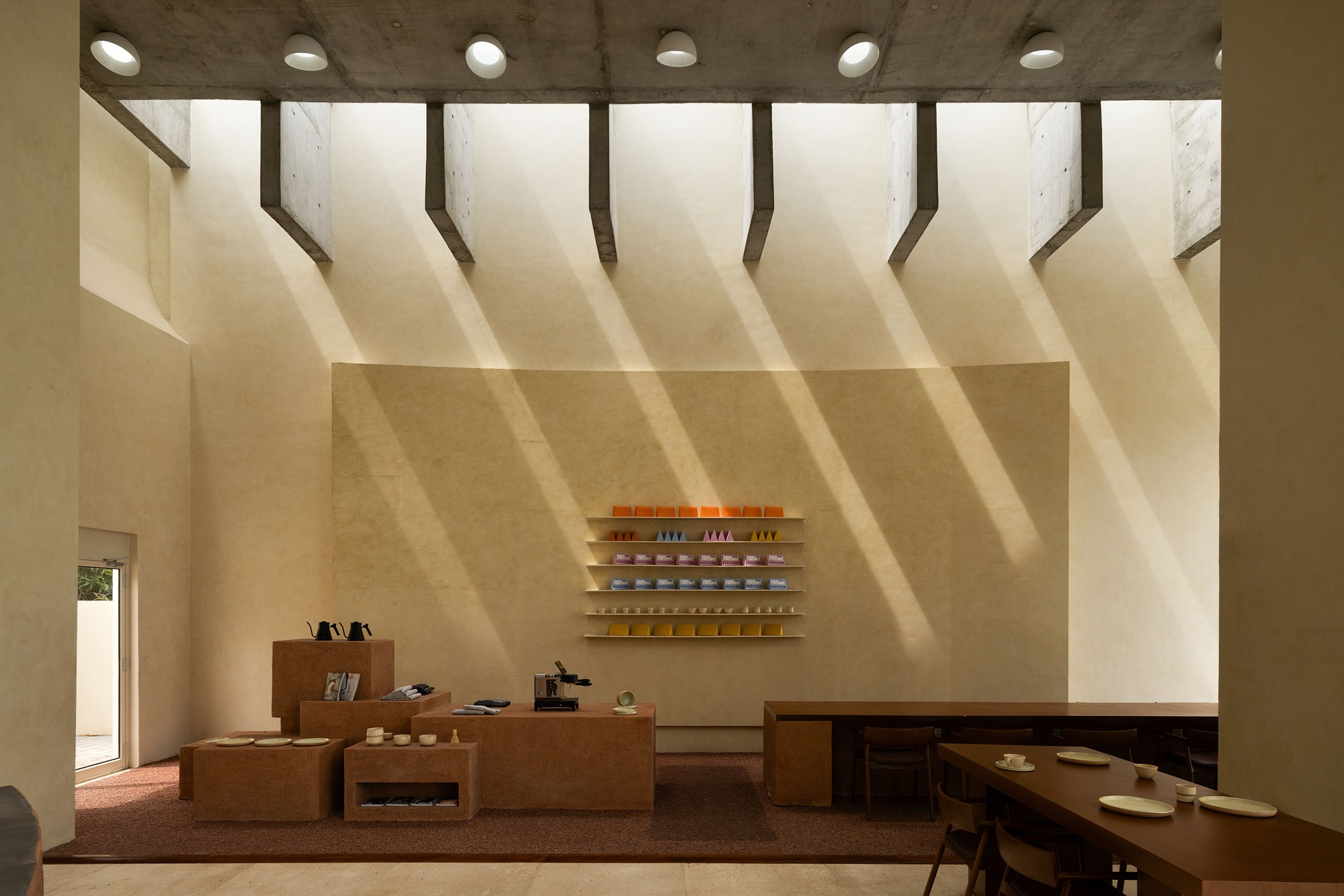 Interior wall with timber shelving and cork display tables illuminated by diagonal shadows from overhead skylights