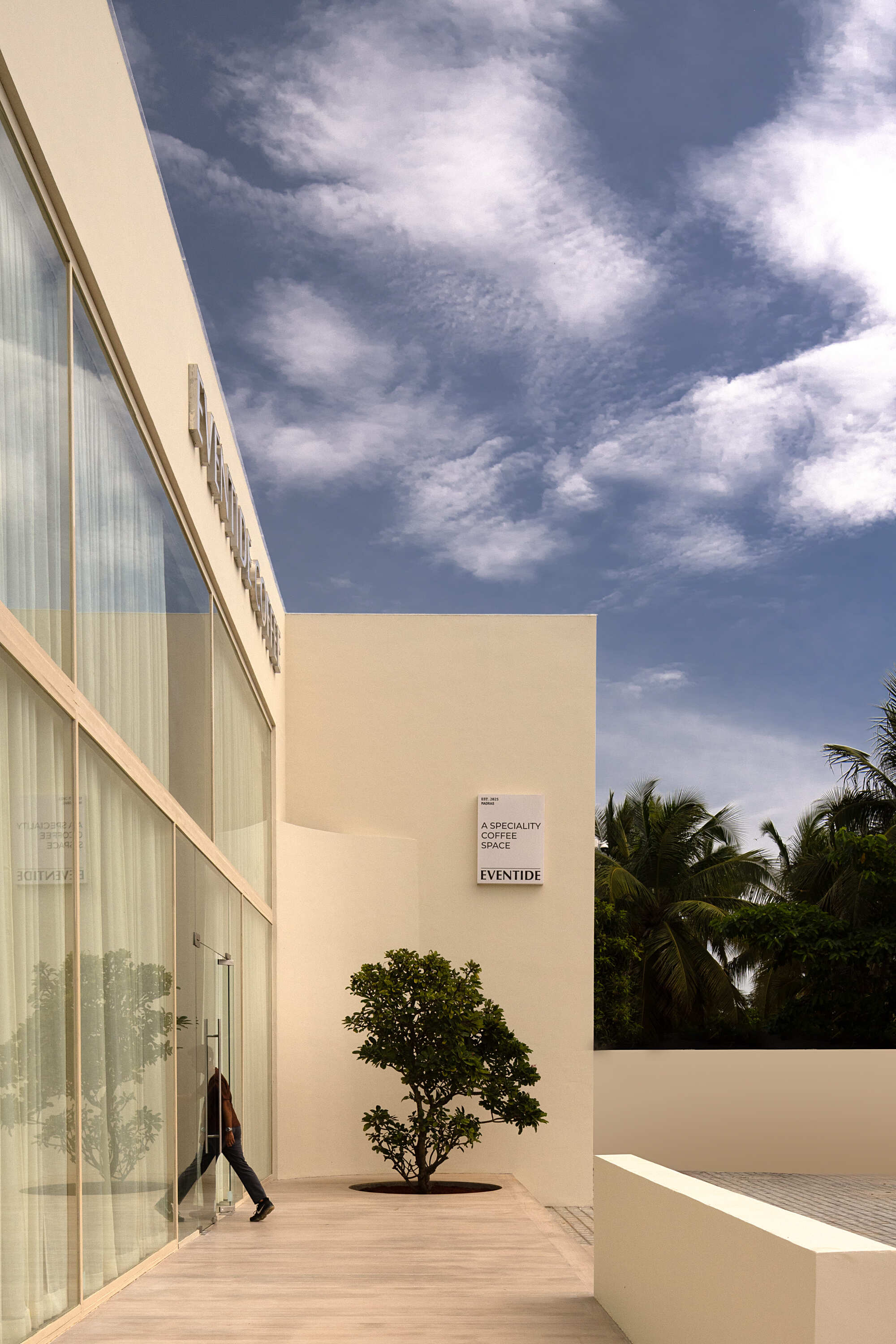 Exterior walkway with translucent glass facade and single tree planted in paved terrace under cloudy sky