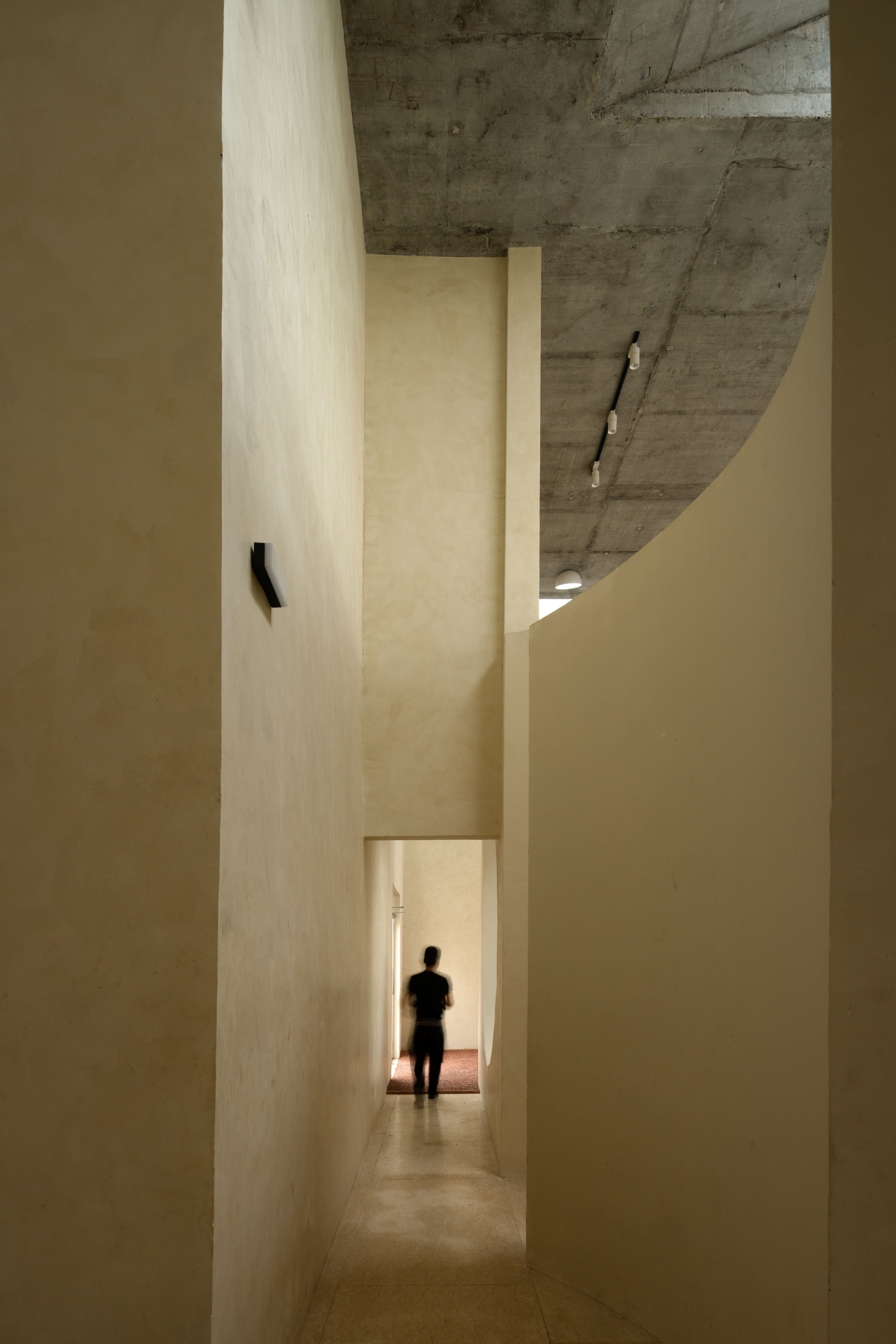 Vertical corridor with plastered walls and exposed concrete ceiling as a figure walks toward light