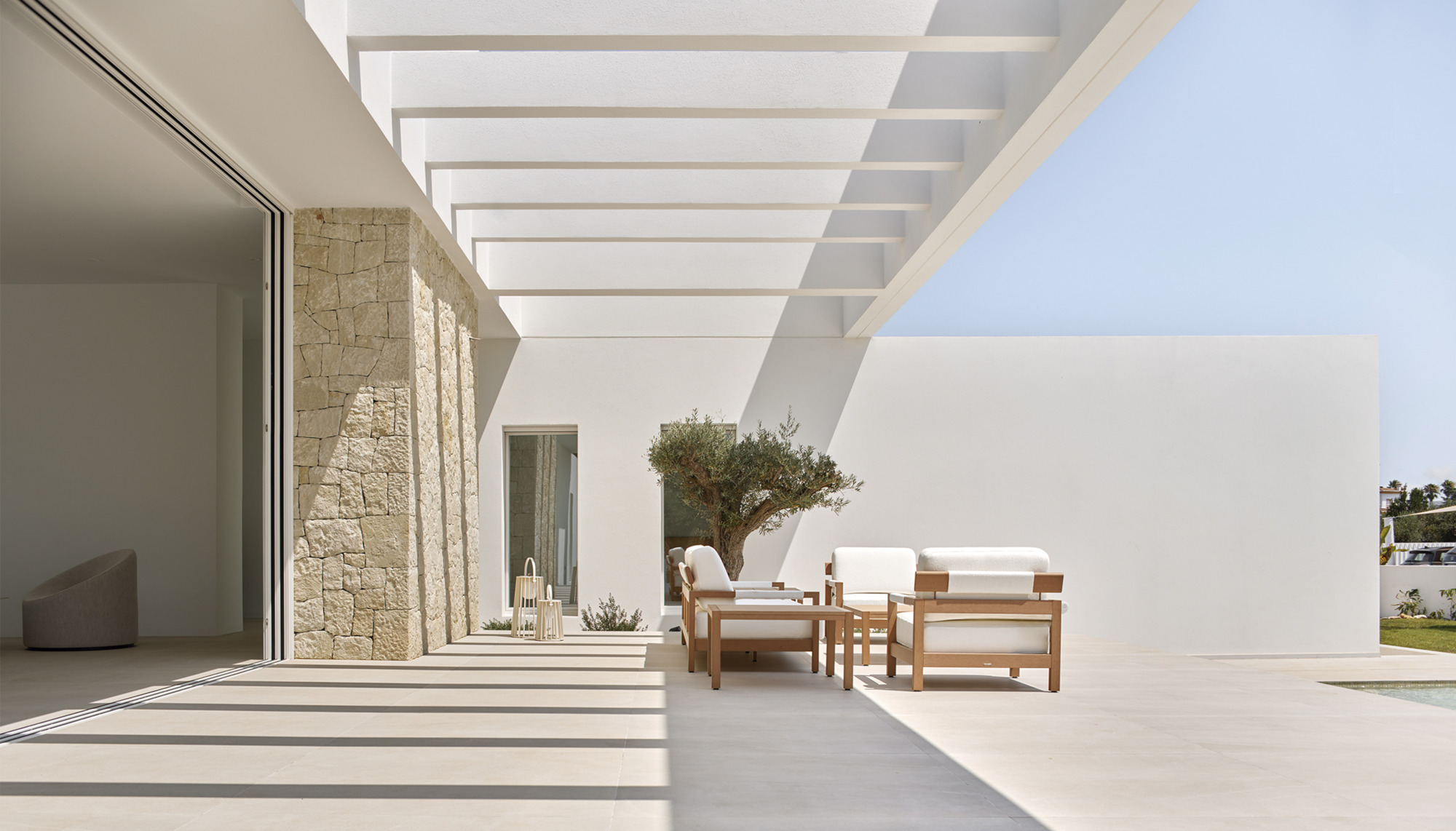 Covered terrace with slatted pergola beams casting shadows over pale stone paving and outdoor furniture