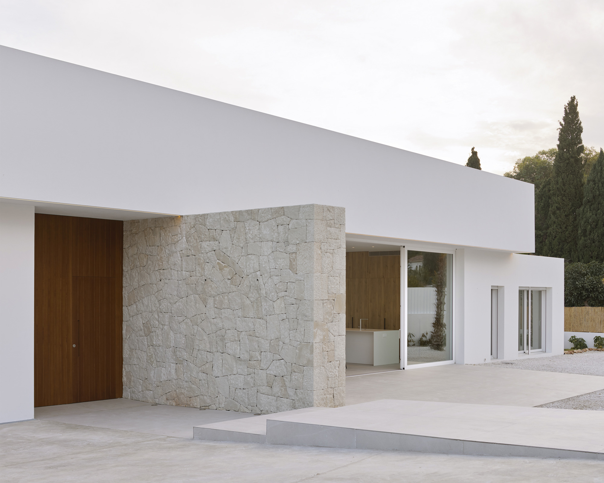 Facade composition of white stucco volumes and limestone cladding with cypress trees visible beyond