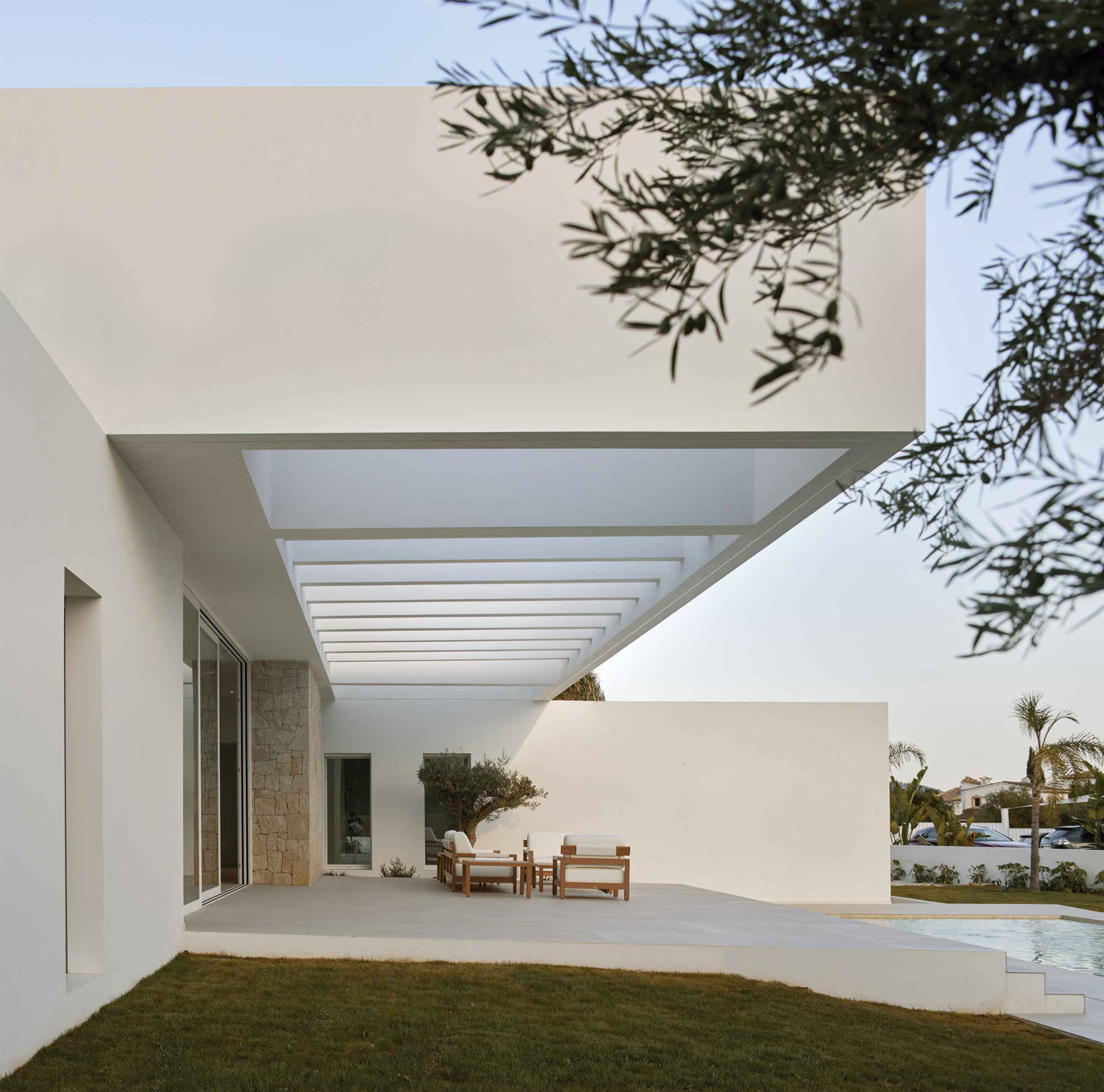Covered terrace with slatted canopy and timber furniture framed by olive branches overhead