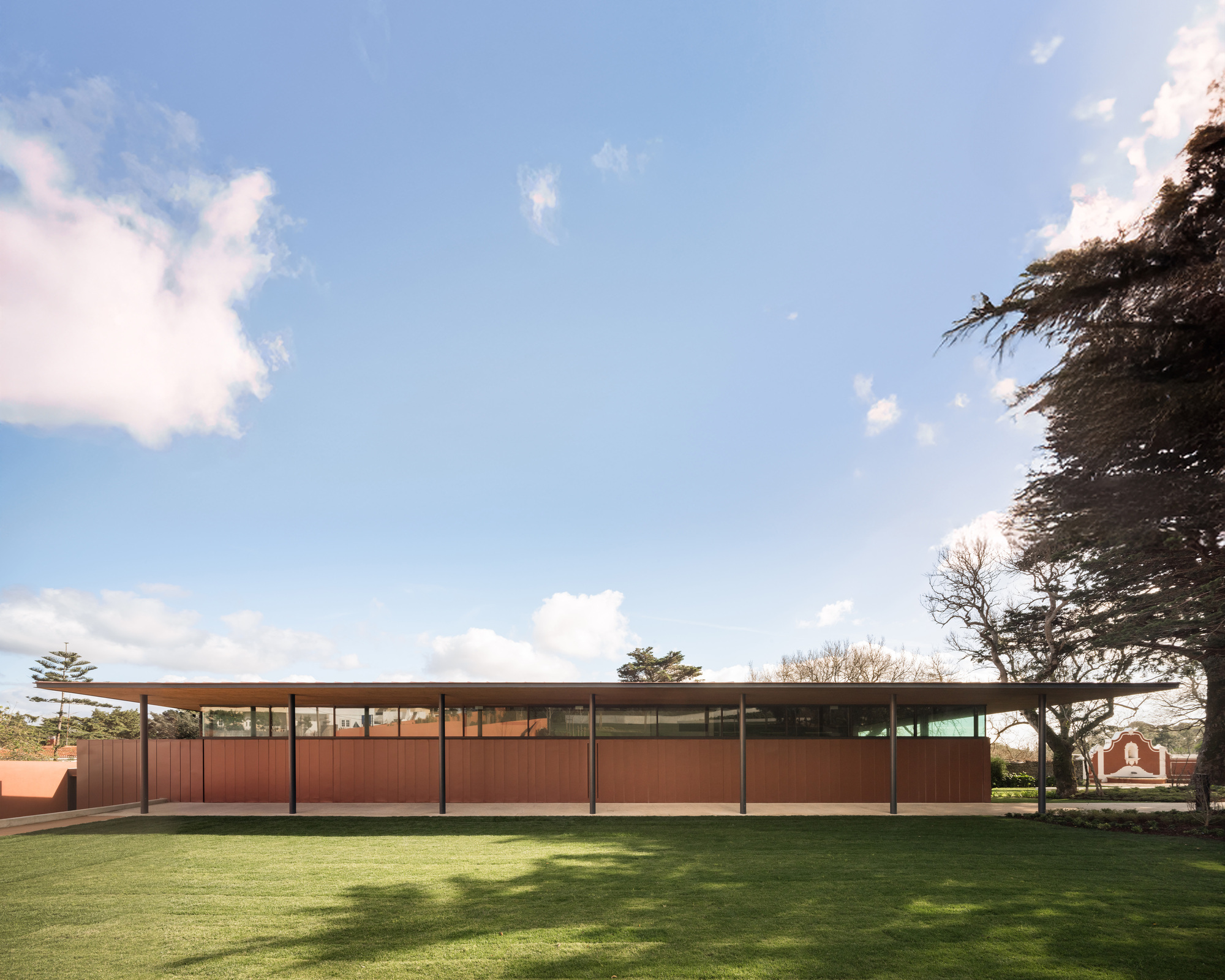 Horizontal pavilion with weathered steel panels and flat roof set in a grass lawn under cloudy skies