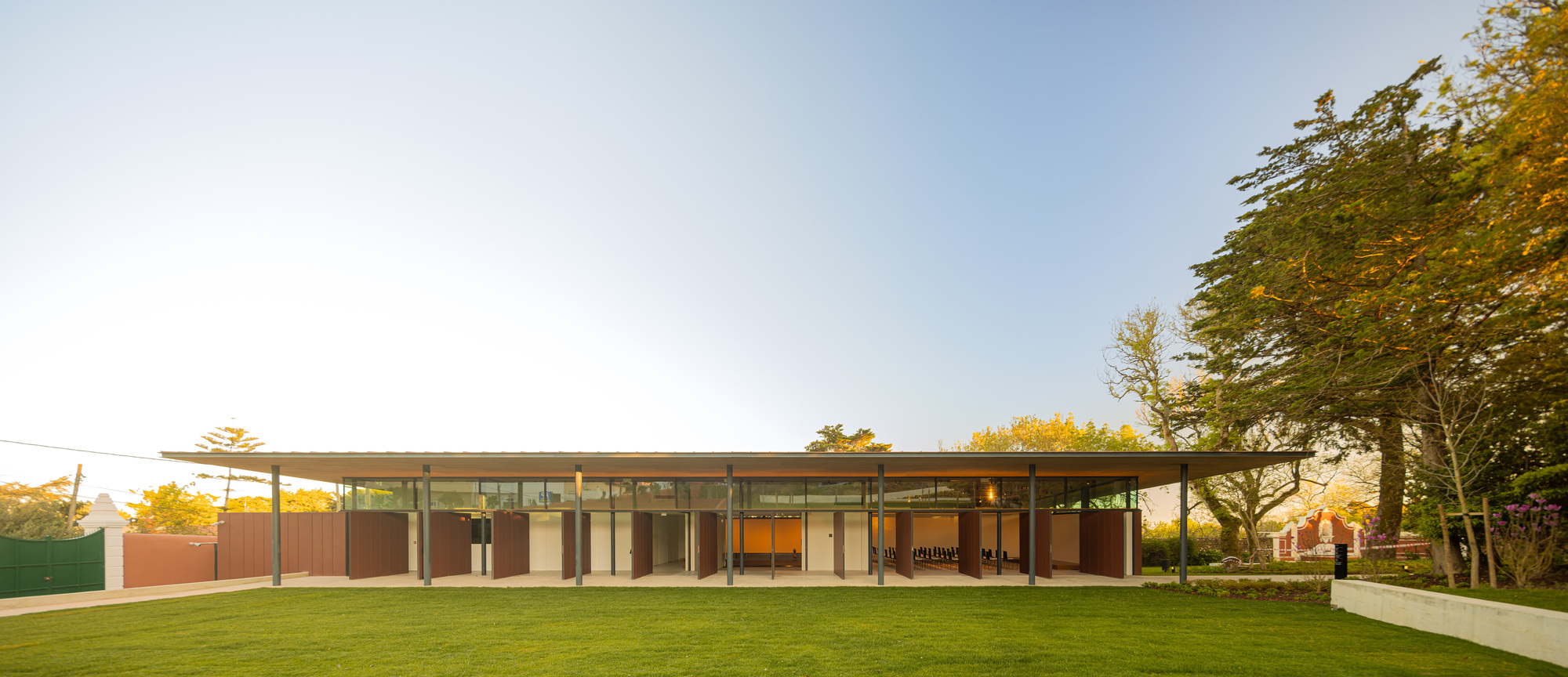 Glass and steel pavilion with overhanging roof on a lawn at golden hour with trees flanking