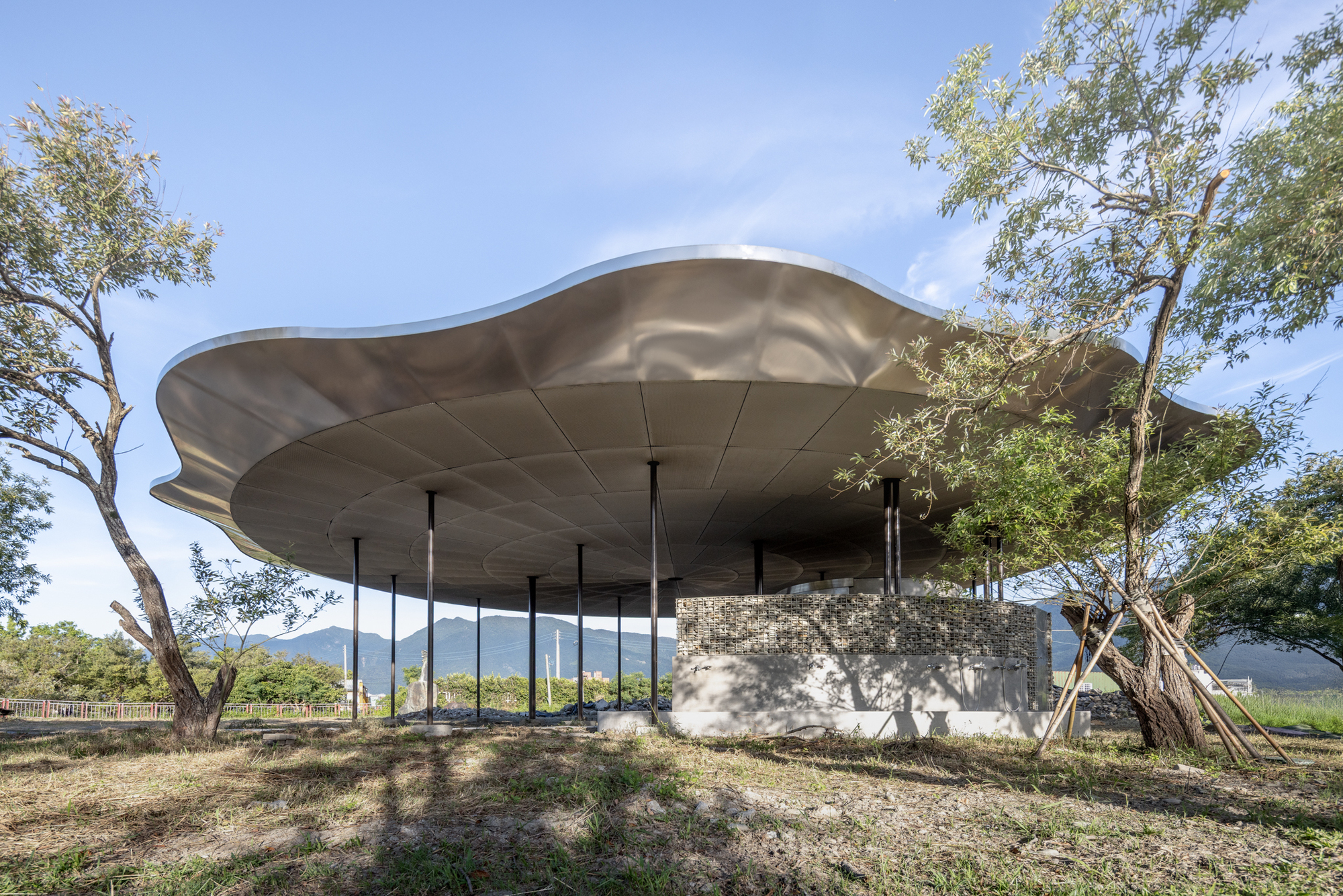 Undulating metal roof canopy supported by slender columns over a stone base surrounded by trees