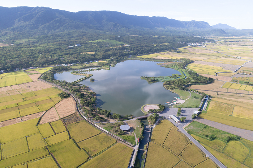 Aerial view of pavilions and curved roofs beside a reservoir surrounded by rice fields and mountains