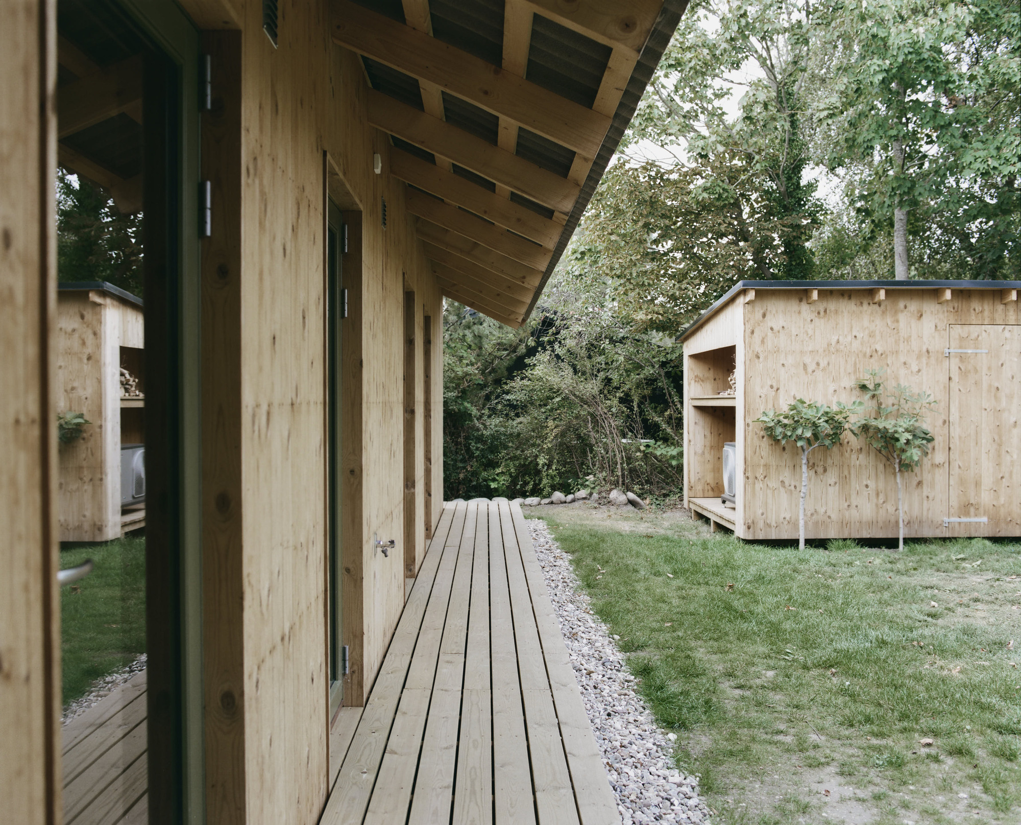 Timber deck connecting two pale wood pavilions beneath a cantilevered eave and surrounding trees