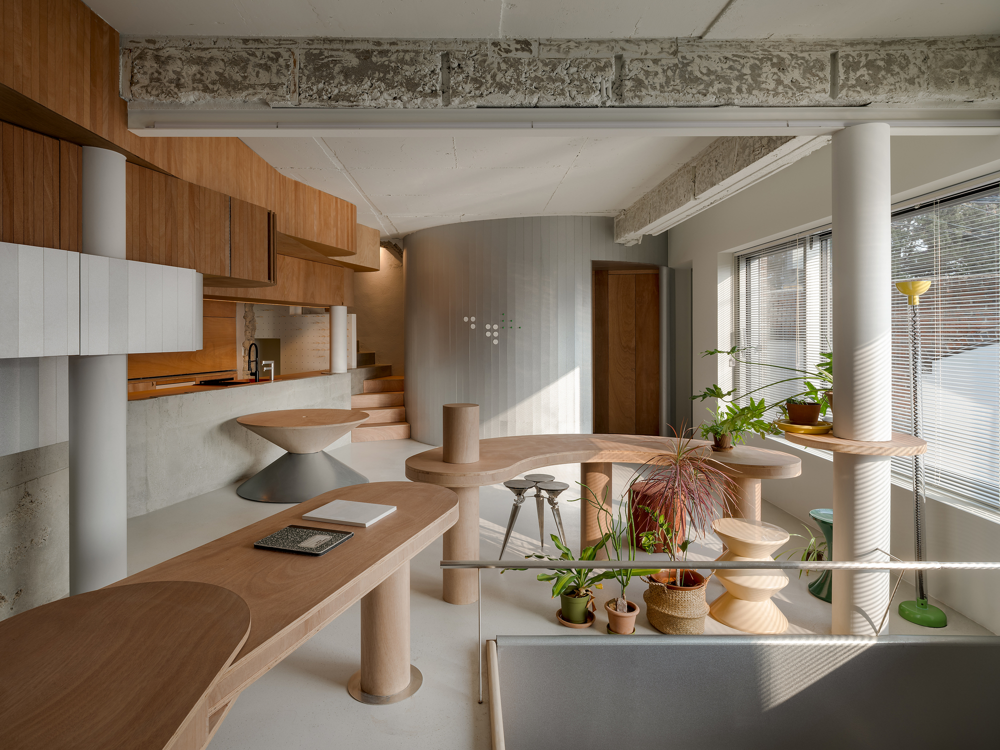 Open interior with sculptural timber table, cylindrical columns, and potted plants beneath exposed concrete beams in afternoon light