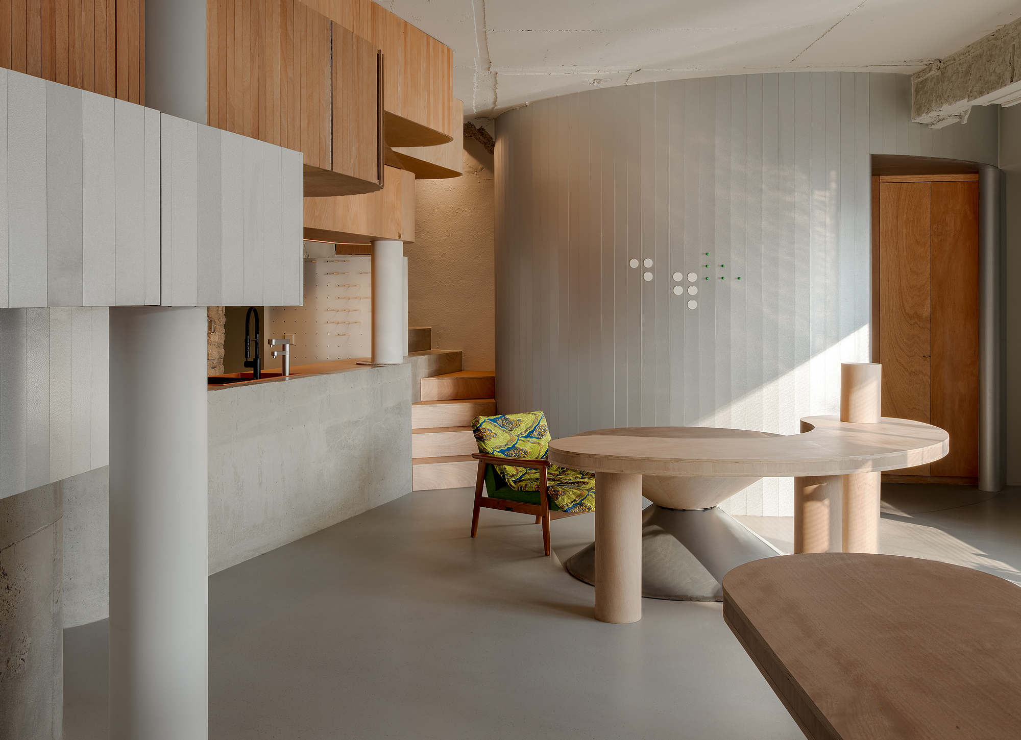 View across the curved timber table toward the sunlit grey wall and wooden staircase rising to the mezzanine