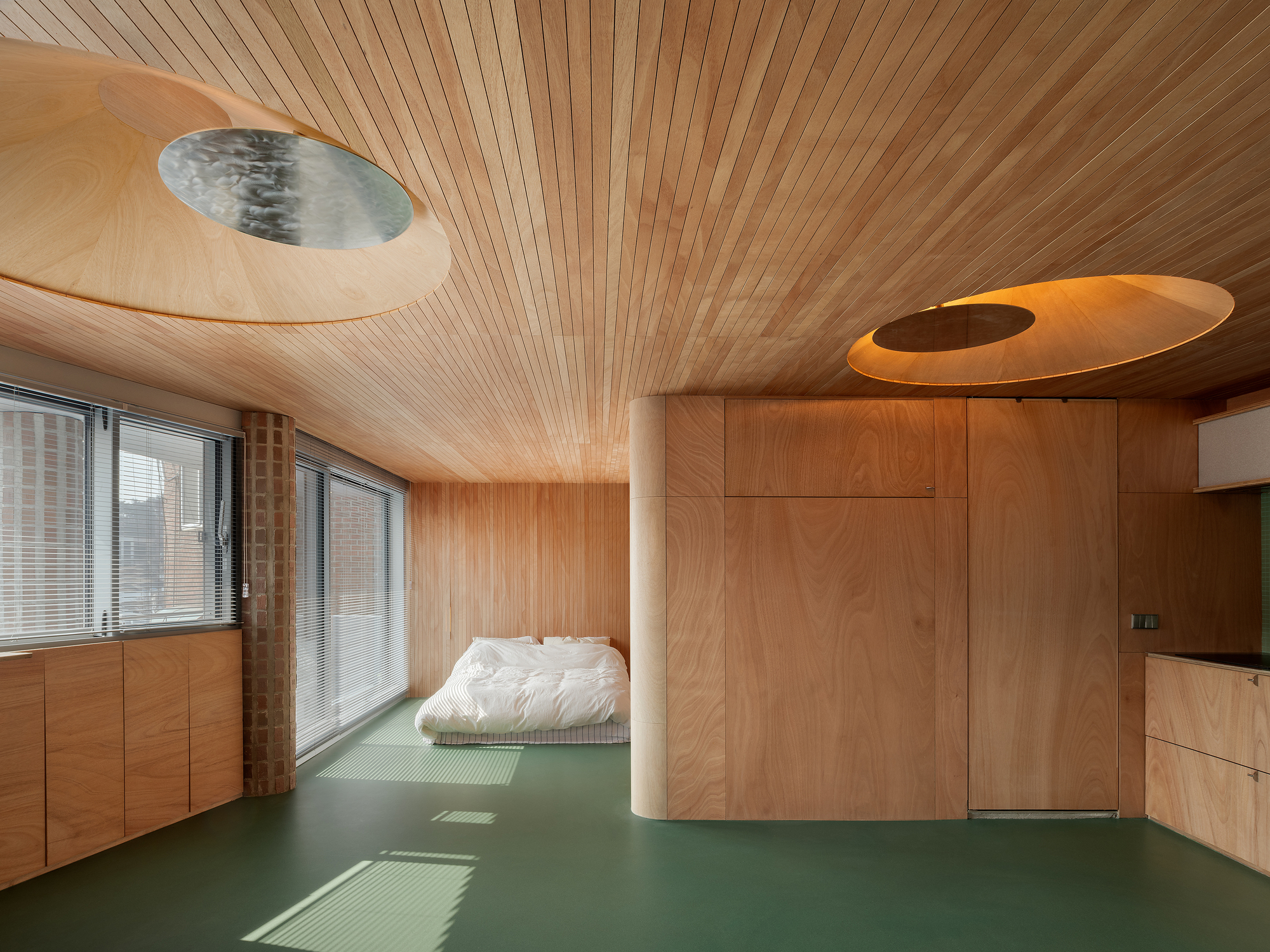 Bedroom with slatted timber ceiling, circular skylights, green floor, and white cylindrical column beside the low bed