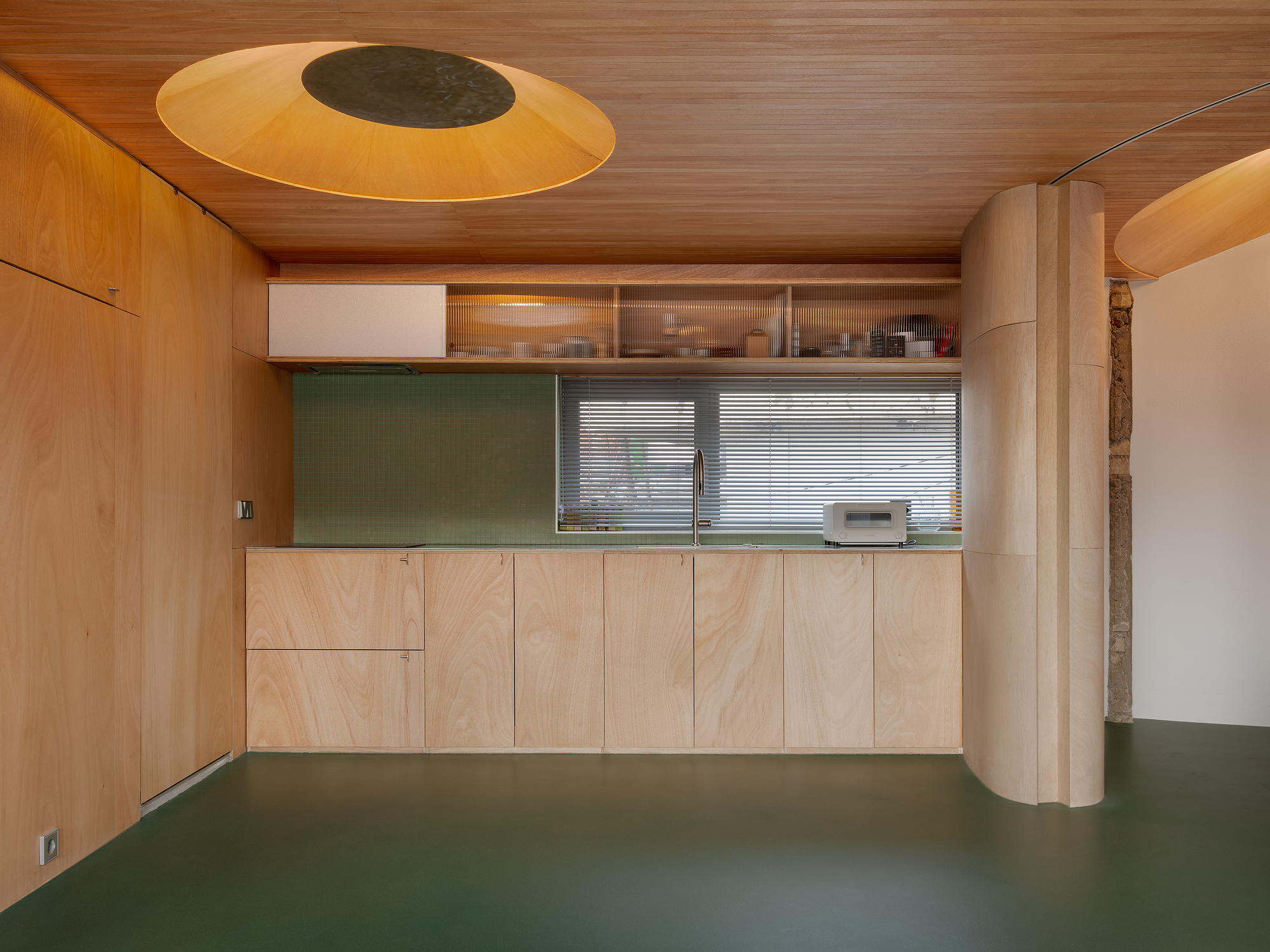 Plywood kitchenette with horizontal window and circular ceiling oculus above green floor