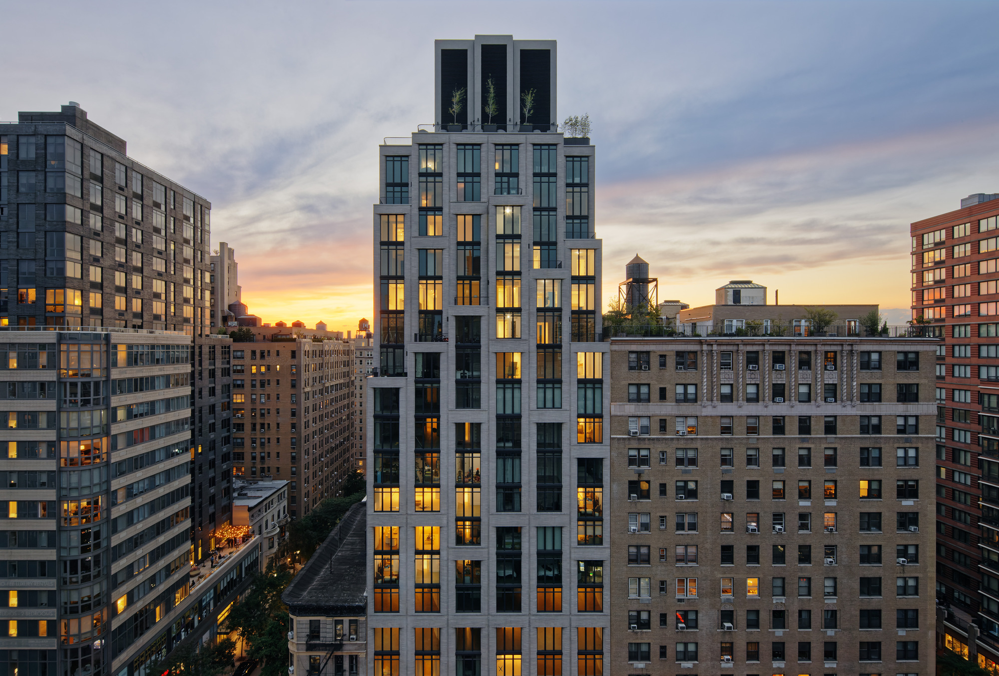 White precast facade with floor-to-ceiling windows lit from within at sunset among neighboring buildings