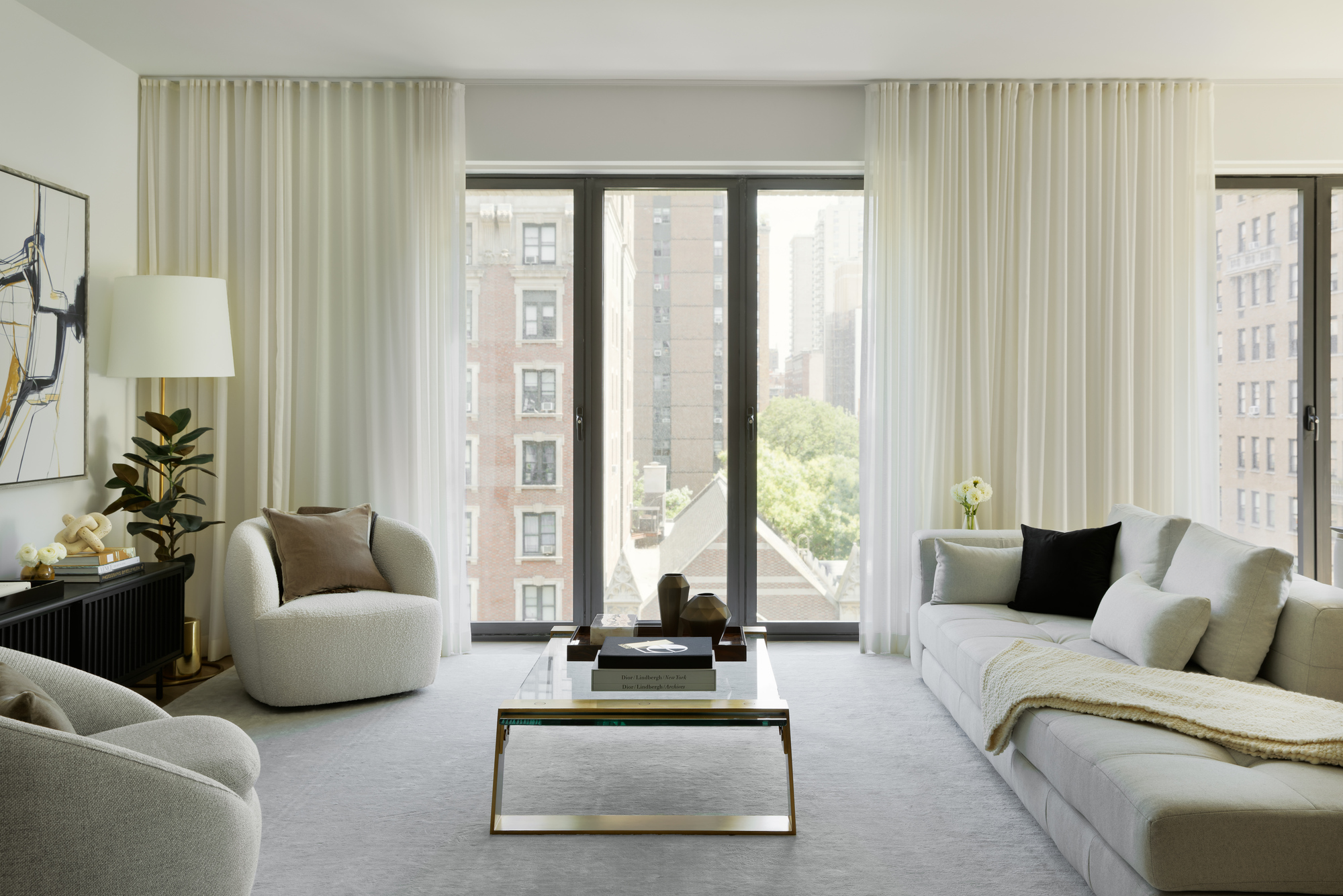 Living room with floor-to-ceiling windows, sheer curtains, and upholstered seating overlooking surrounding buildings