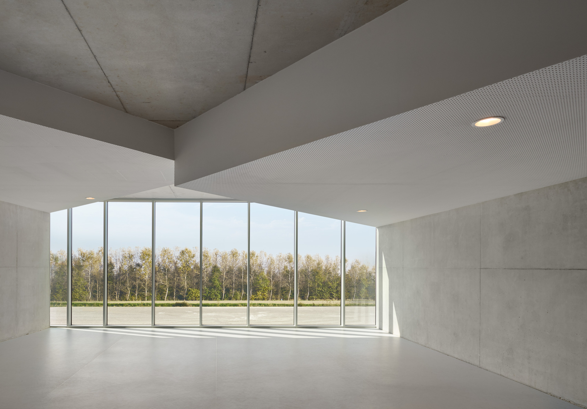 Interior room with concrete walls and ribbed ceiling opening to glazed facade overlooking trees