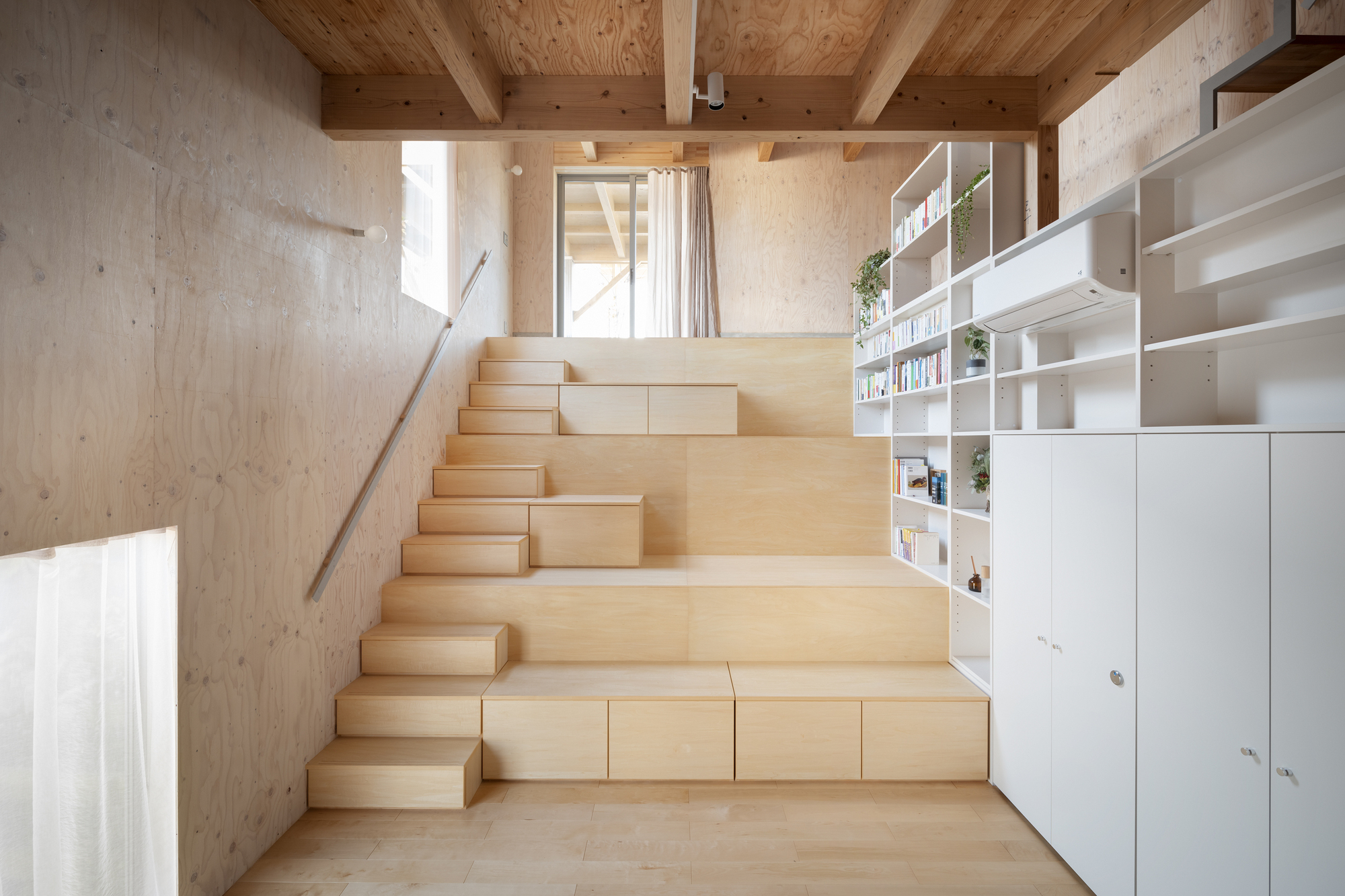 Wide plywood steps with pull-out storage drawers facing white shelving under timber beam ceiling