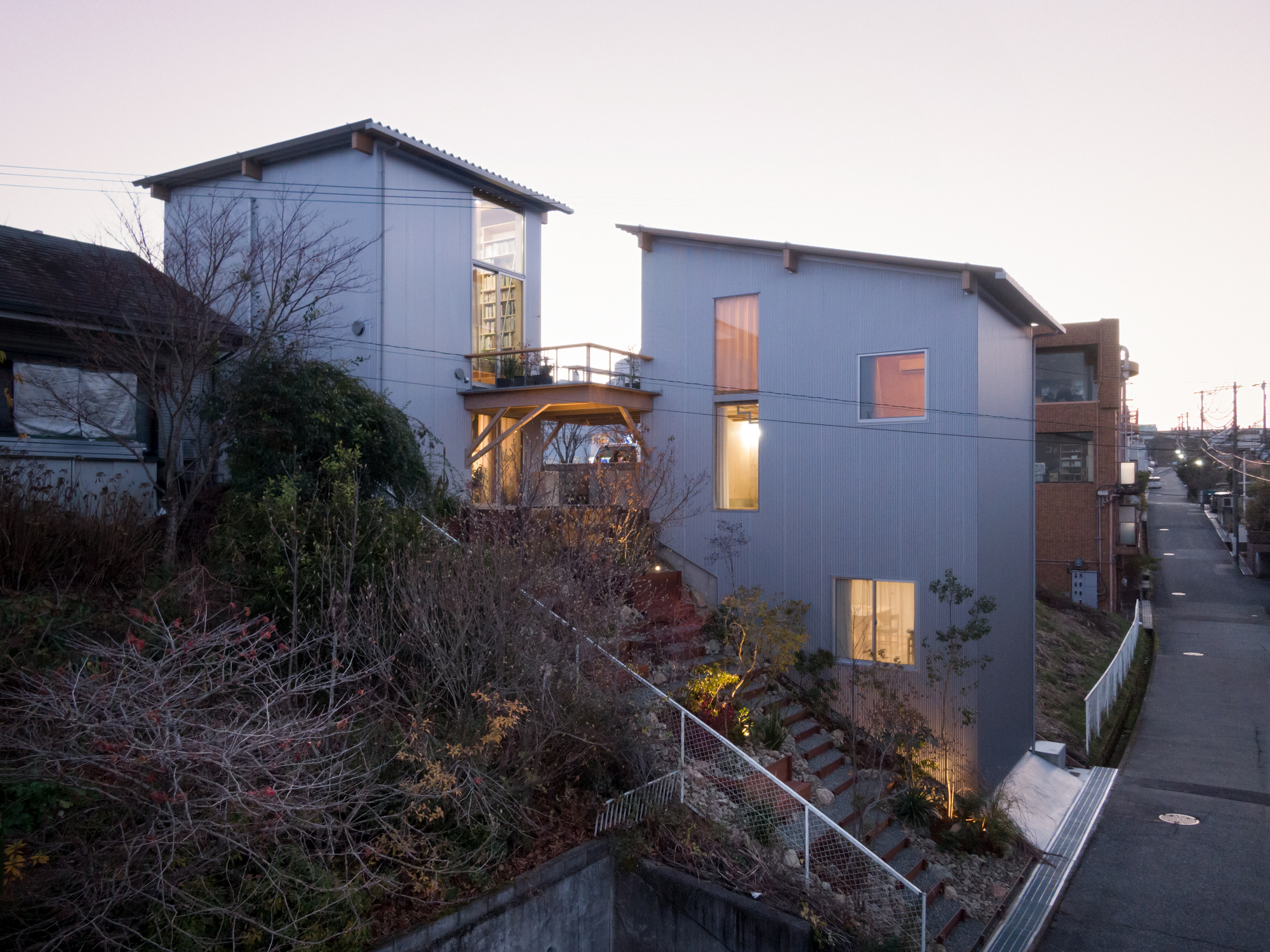 Hillside facade with corrugated metal cladding and illuminated windows at dusk overlooking a sloped landscape