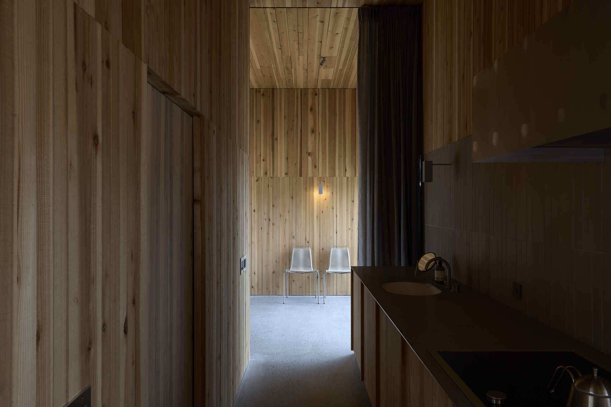 Timber-lined corridor with pendant light leading to two white chairs in adjacent room