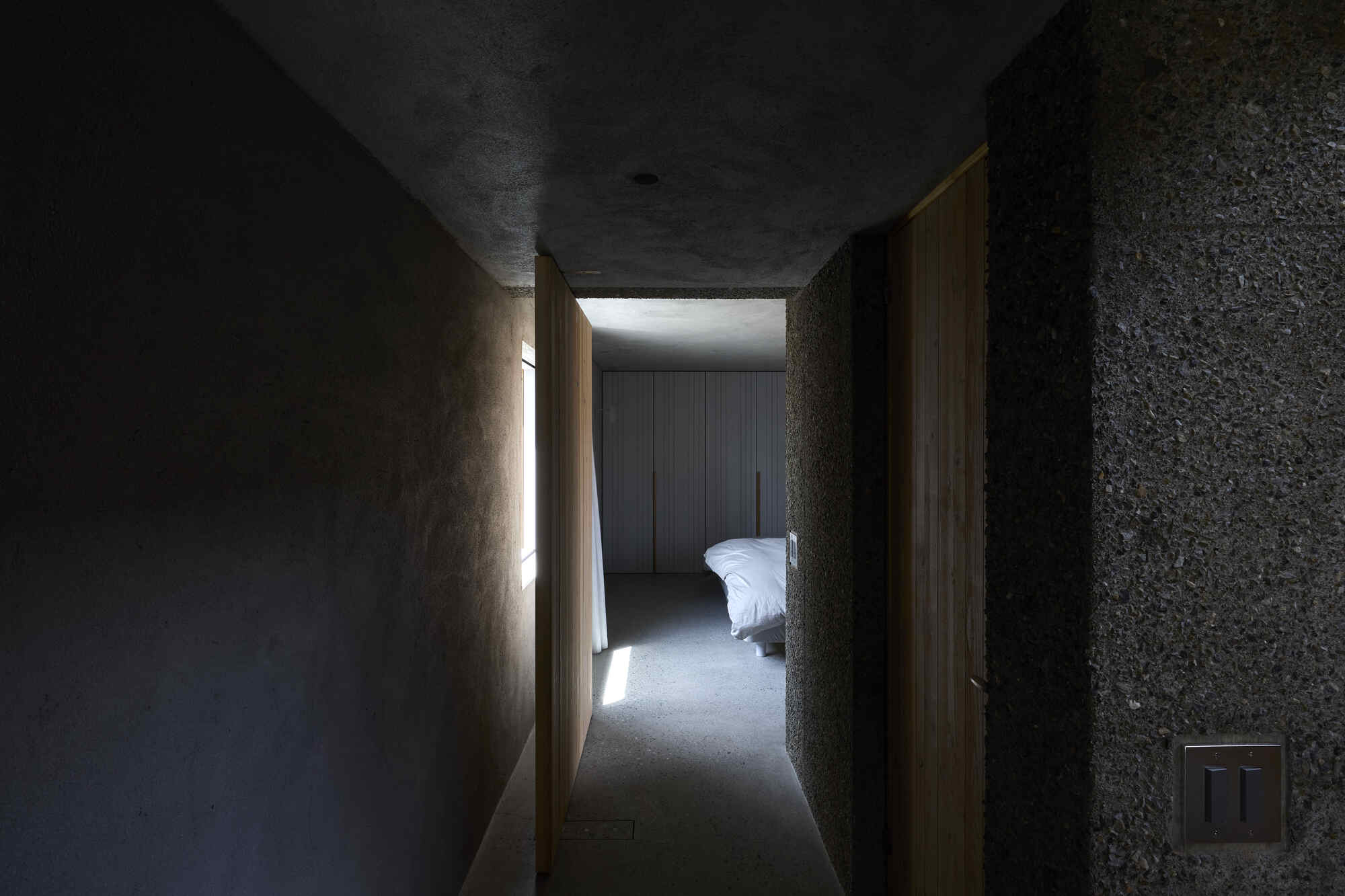 Narrow passage with textured concrete walls and timber door framing bedroom with clerestory light