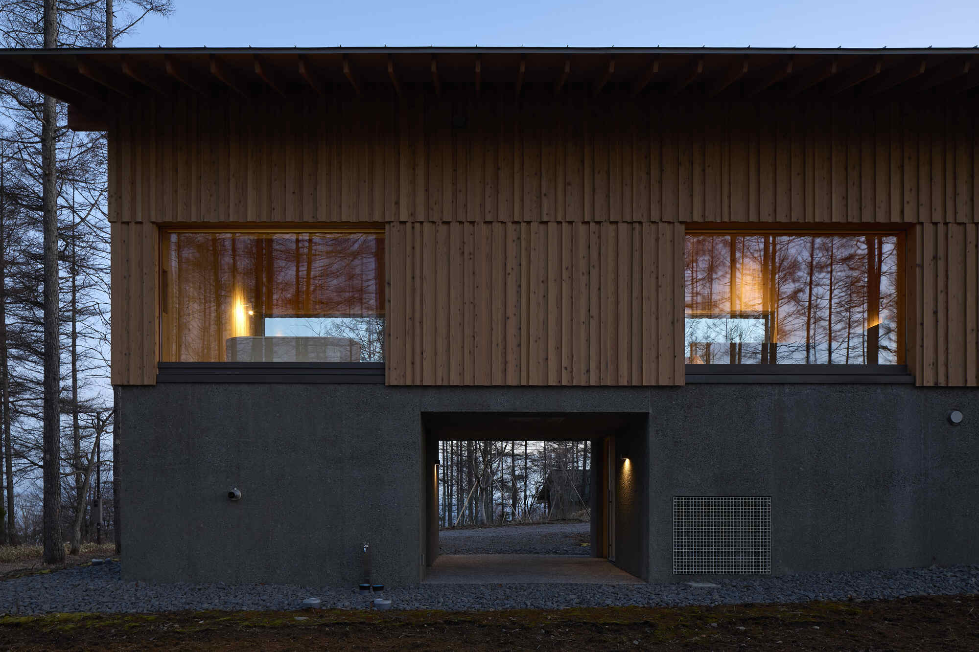 Two-story facade with vertical timber cladding over dark plaster base and covered passageway at dusk