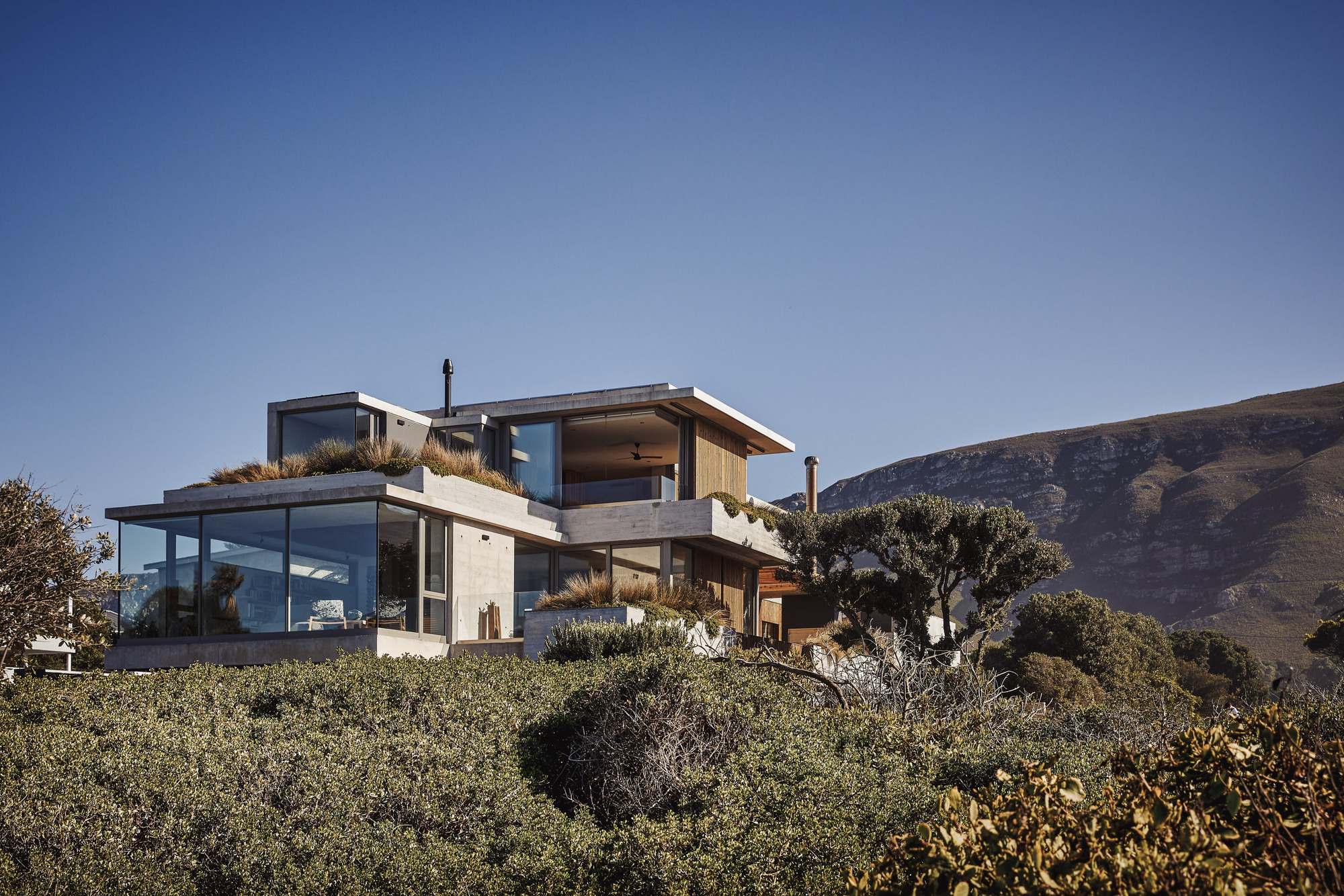 Terraced residence with concrete and timber volumes nestled into a vegetated hillside below distant mountains