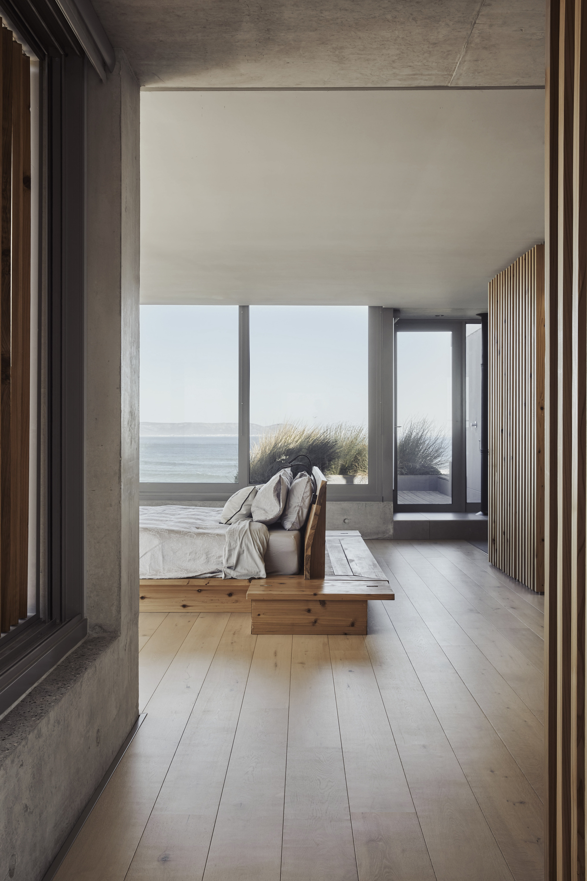 Corridor with timber platform bed and vertical slatted wall looking toward ocean-facing glazing and dune grass