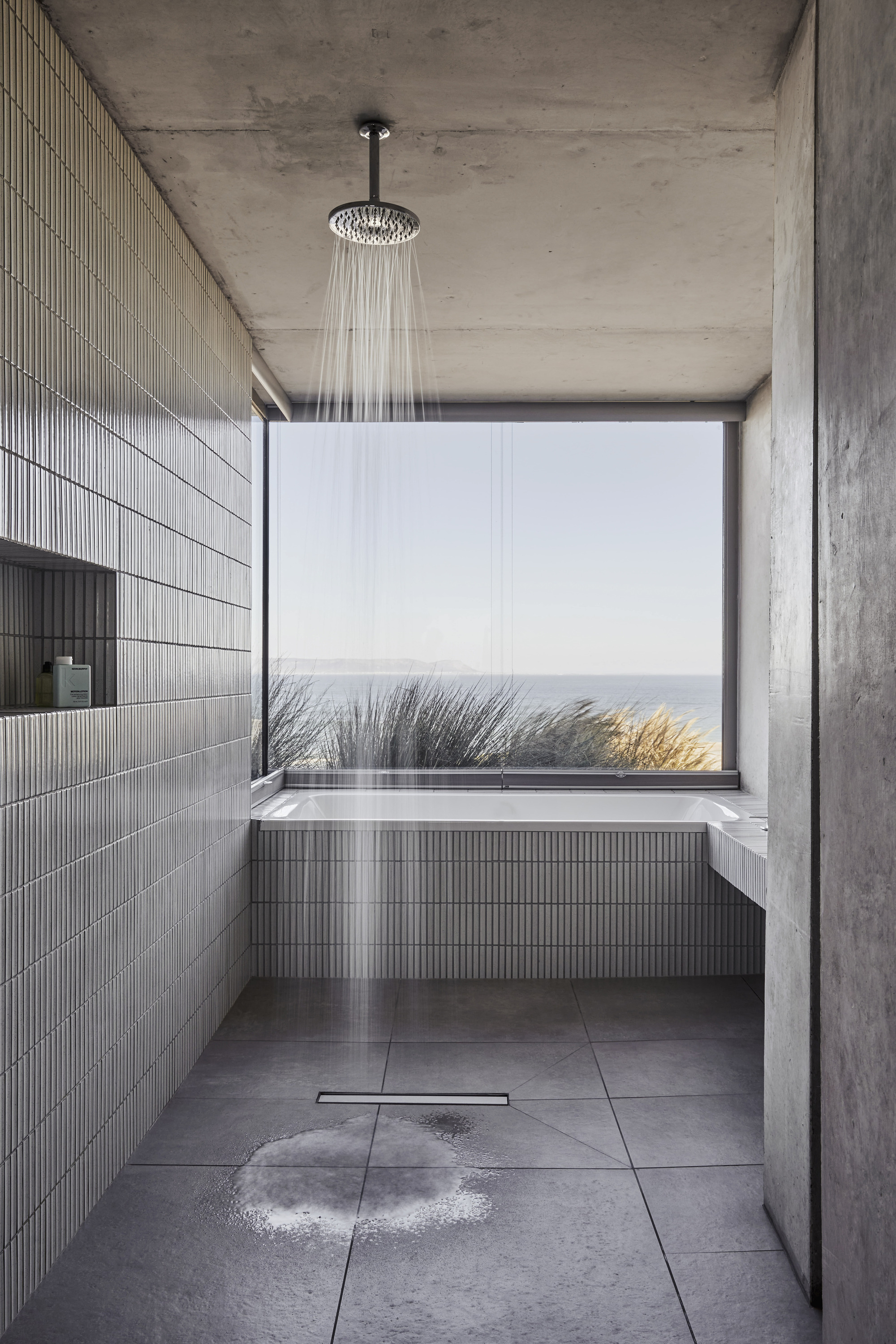 Concrete bathroom with rainfall shower, ribbed tile and picture window framing coastal grasses and ocean