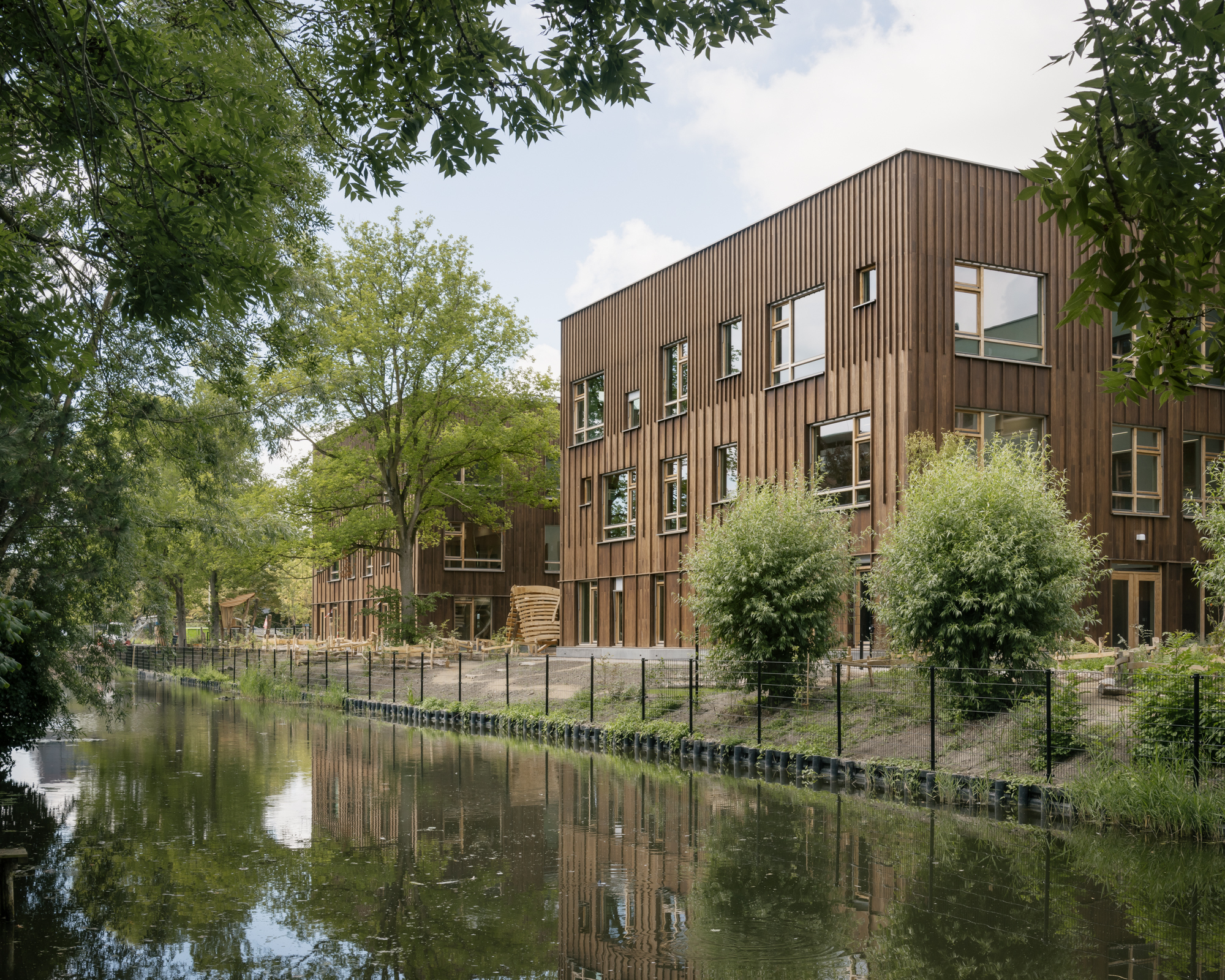 Vertical timber cladding facade with scattered window openings reflected in the adjacent canal