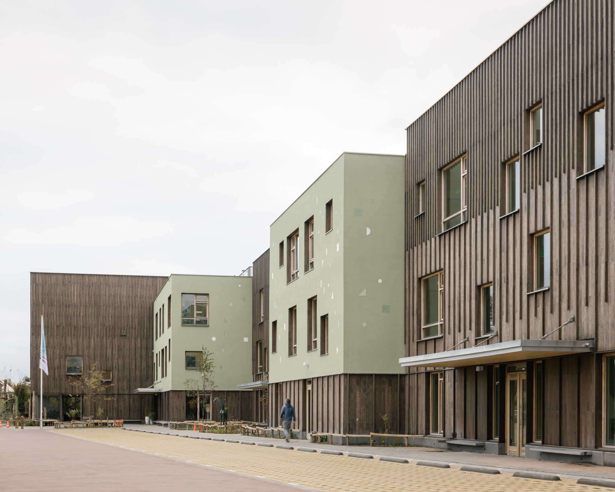 Street view showing the junction of pale green rendered volumes and vertical timber-clad blocks under overcast sky