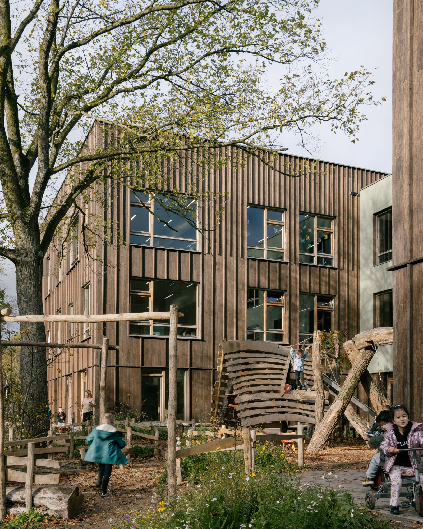 Timber facade with scattered windows viewed through a natural play area with children and a bare tree in foreground