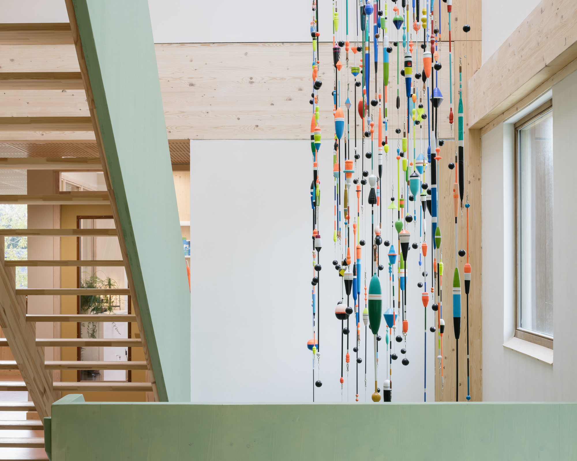 Double-height timber stairwell with painted green guardrail and hanging installation of colorful fishing floats