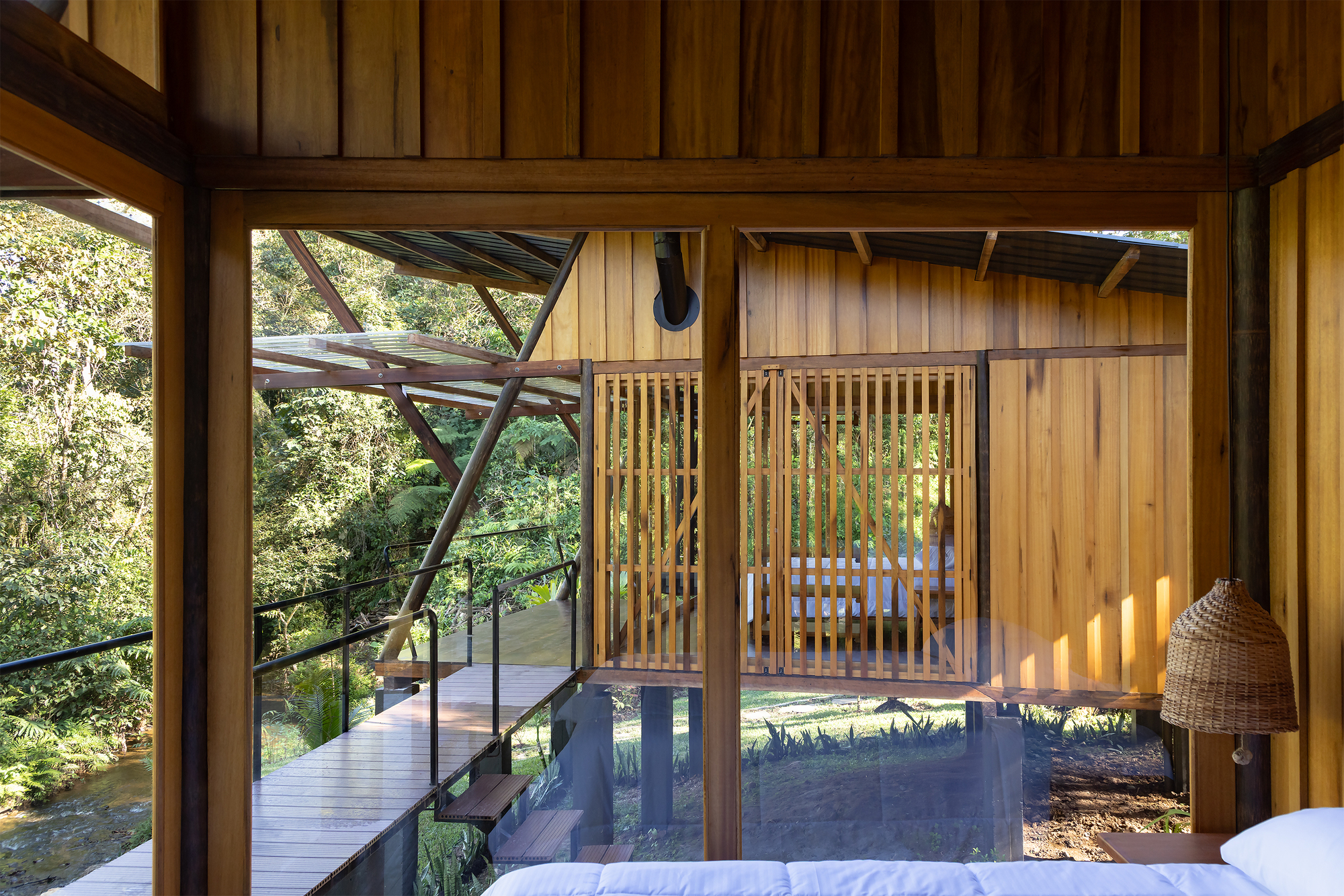View from bedroom through timber-framed glazing to exterior walkway with diagonal bracing and forest beyond
