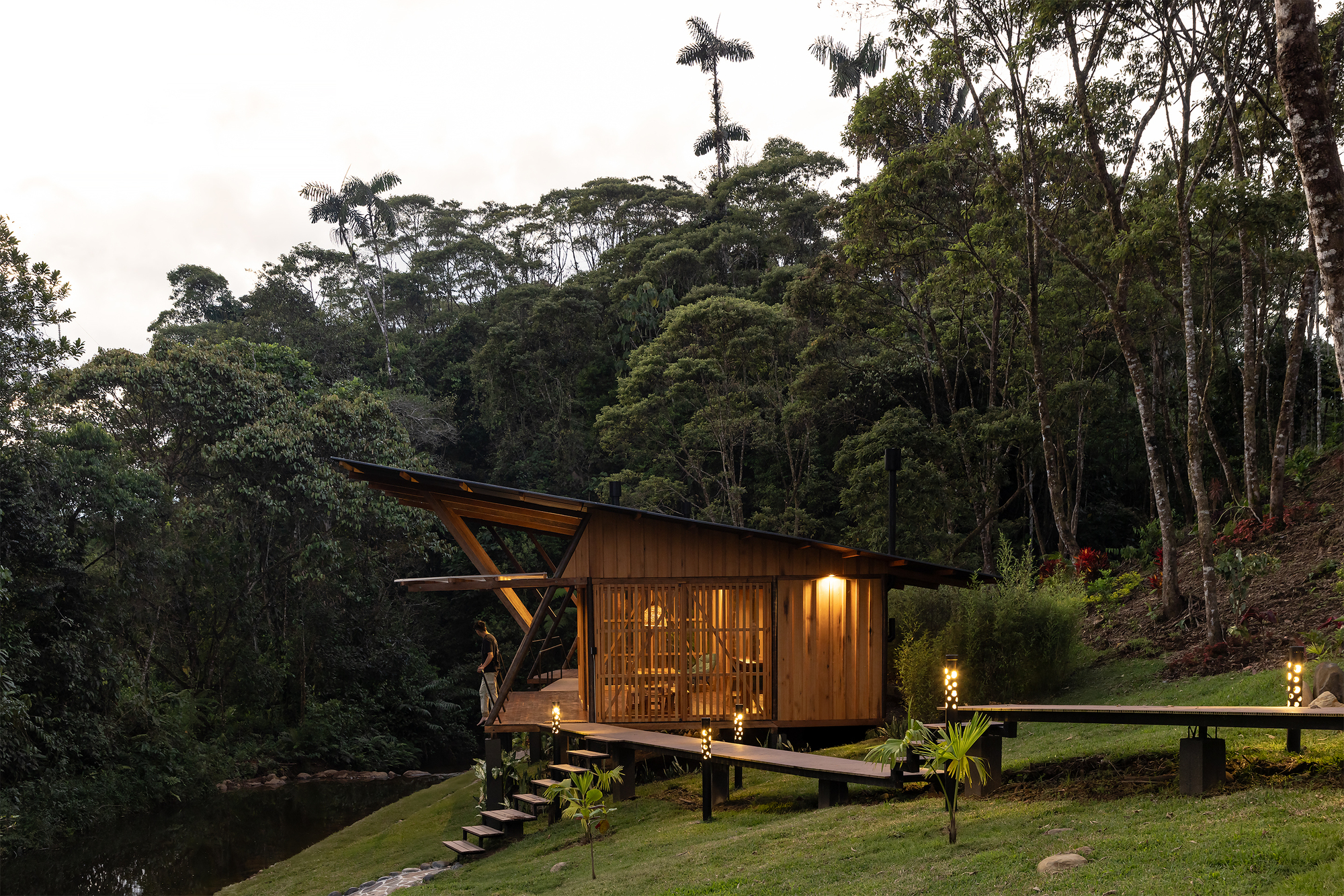 Elevated timber pavilion on pilotis with folded roof and screened facade at dusk beside a pond
