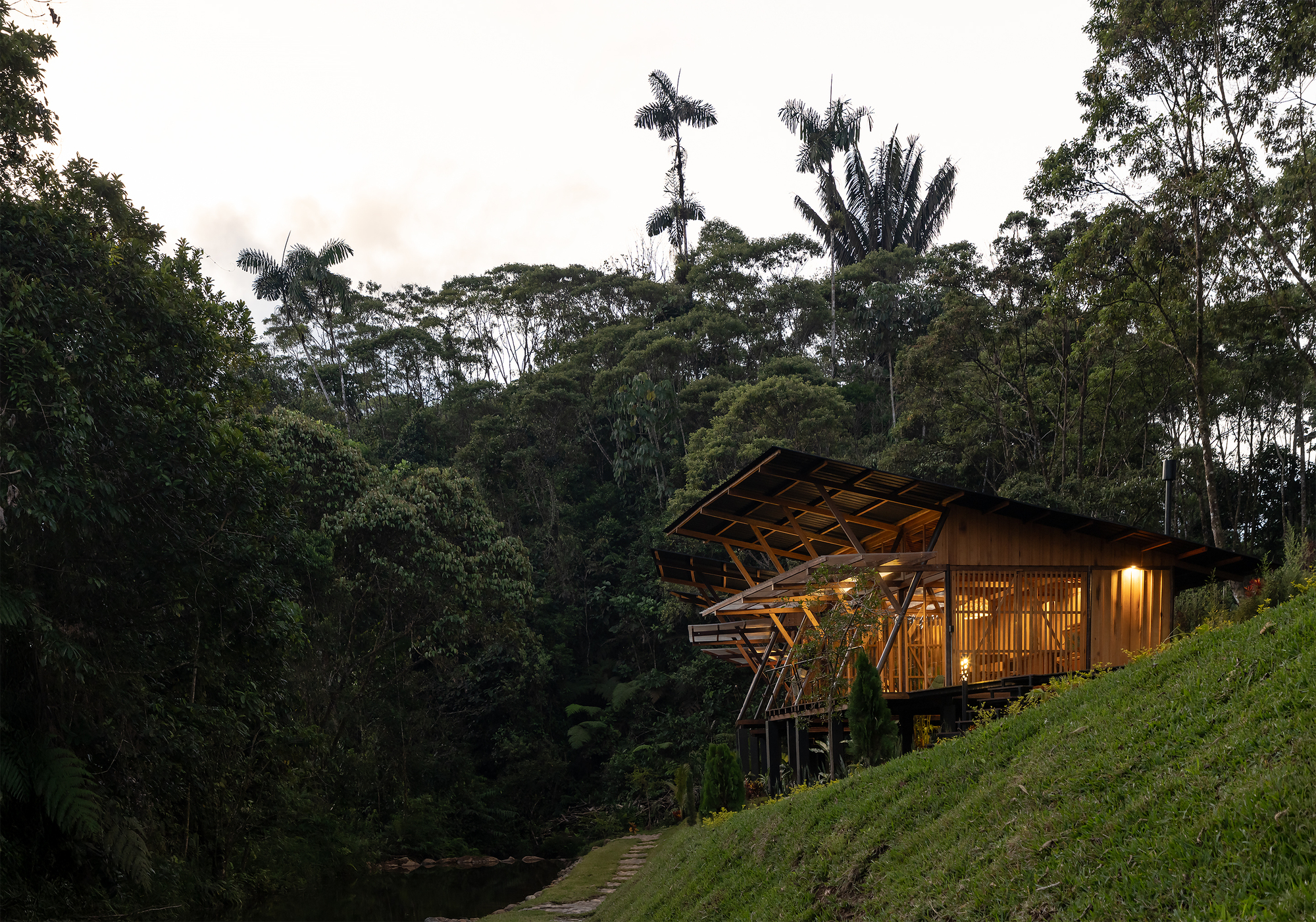 Timber structure with angled roof and latticed facade nestled into hillside among dense tropical vegetation at twilight
