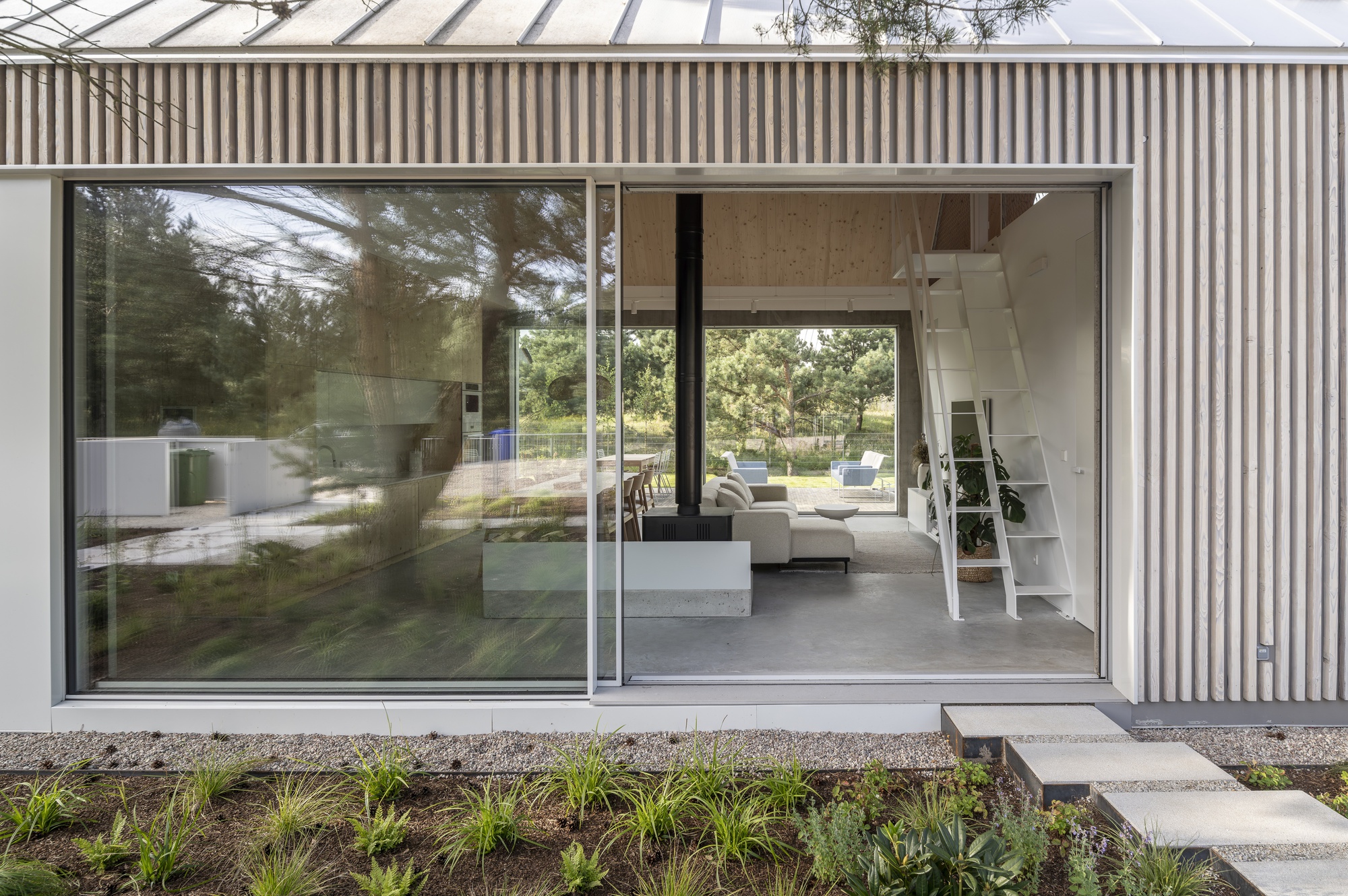 Floor-to-ceiling glazed opening in corrugated metal wall revealing interior living space and garden beyond