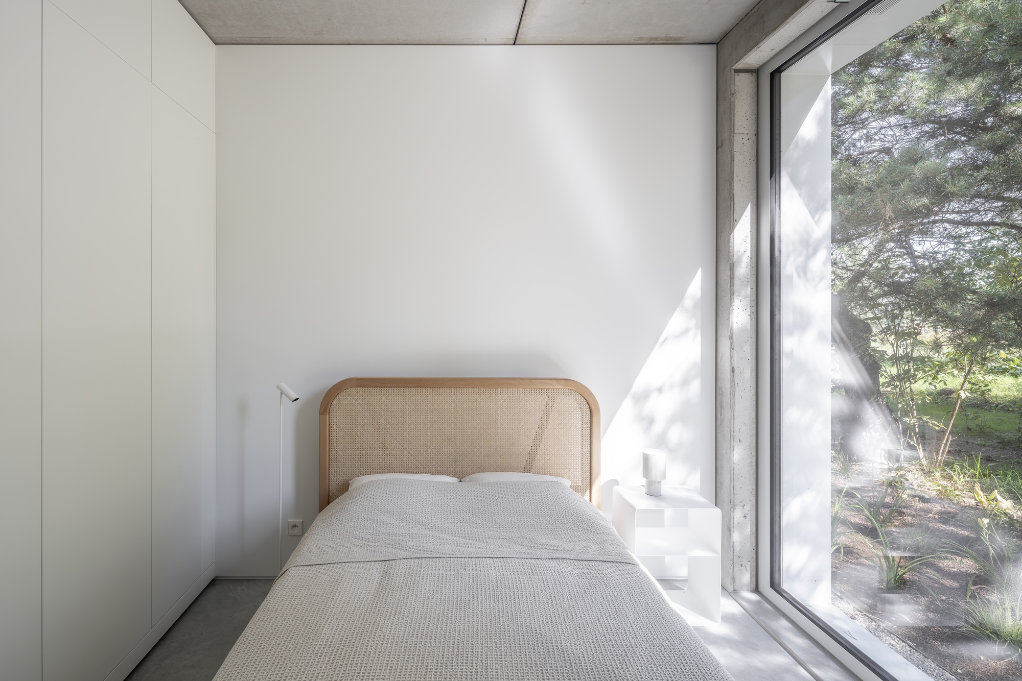 Bedroom with floor-to-ceiling glazing casting dappled sunlight across the white walls and concrete ceiling