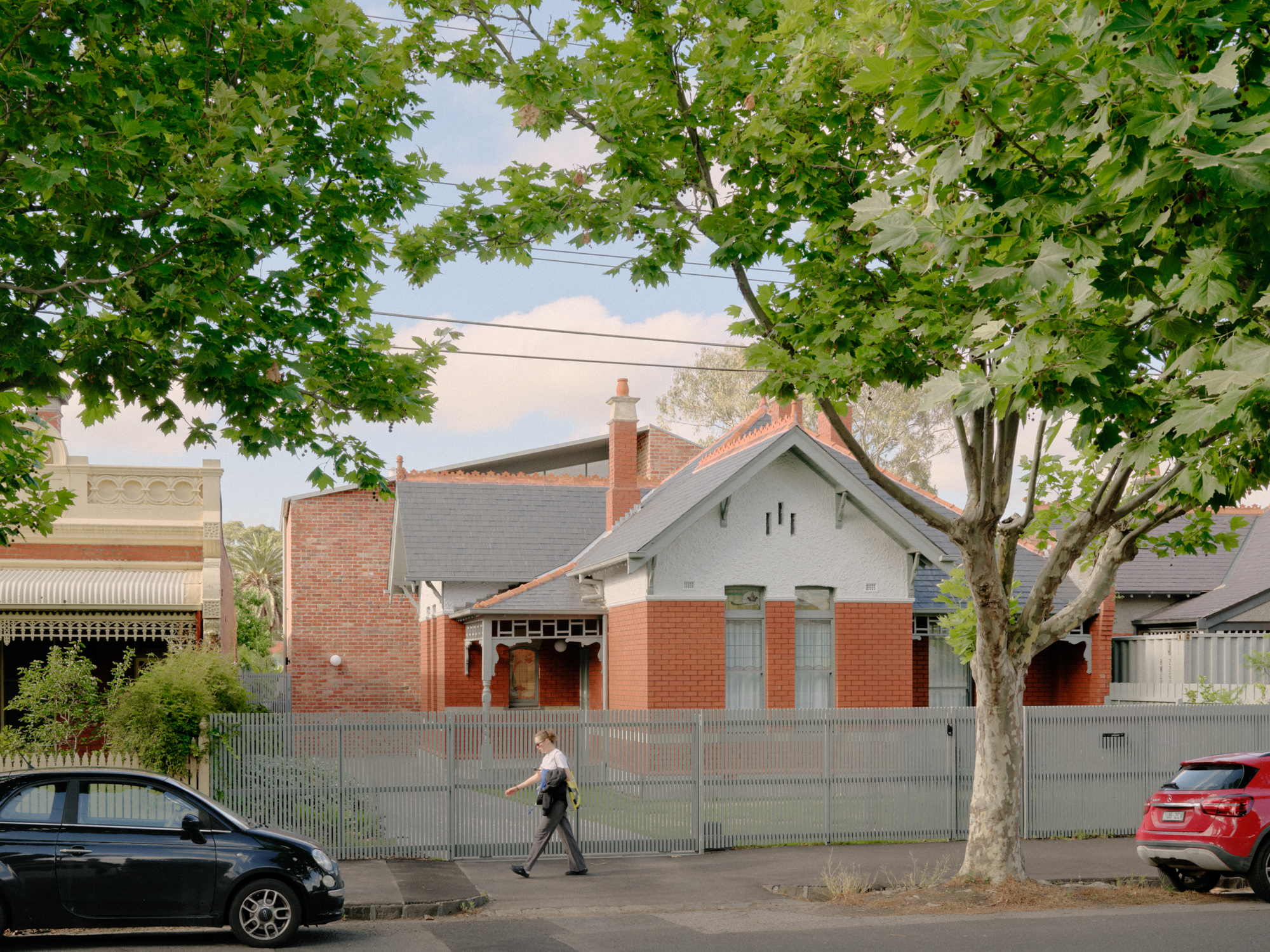 Street view of a gabled house with red brick base and grey painted timber under green tree canopy